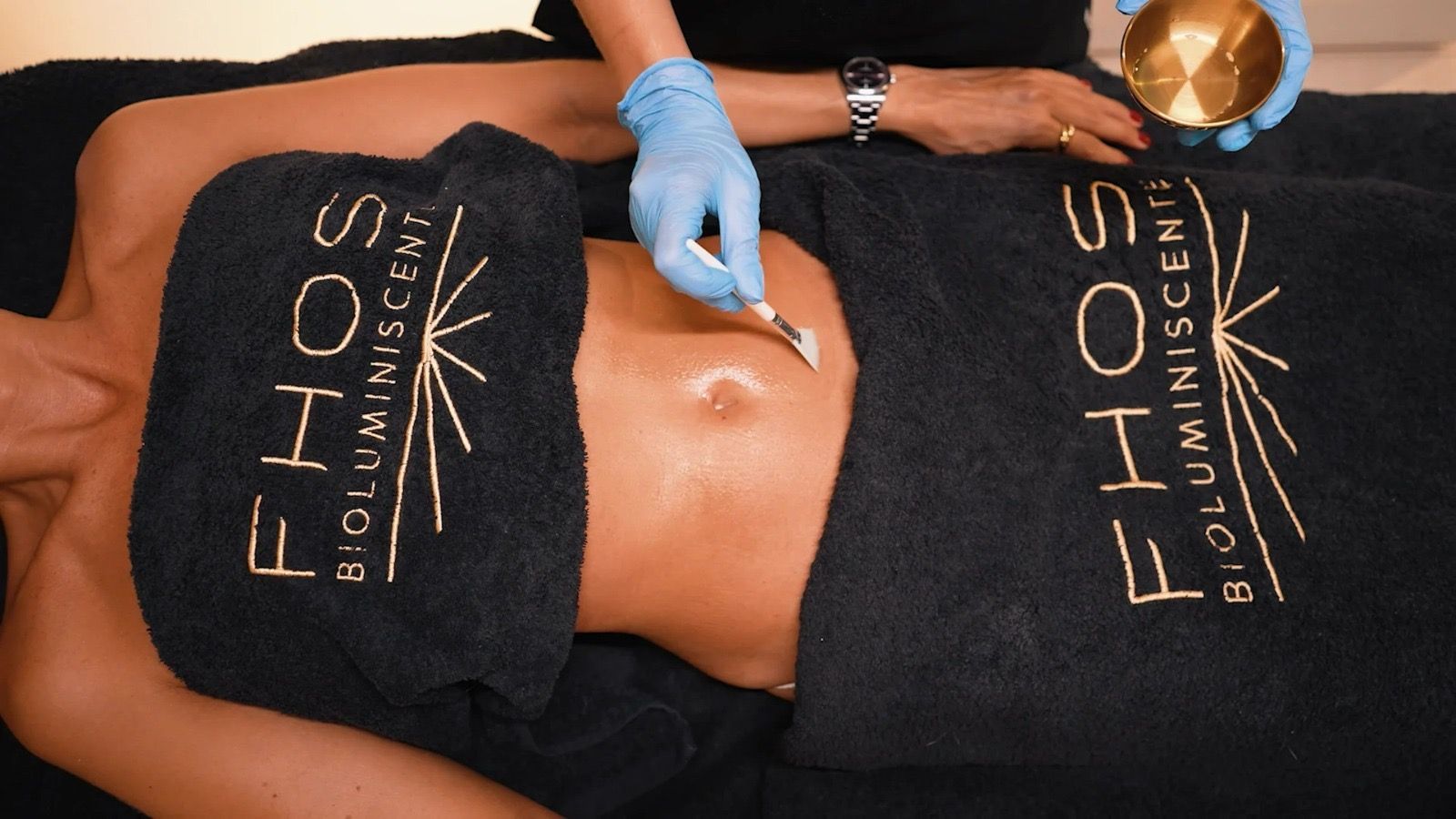 Mujer recibiendo tratamiento abdominal; aplicando producto. Toallas negras con logo dorado. Guantes azules.