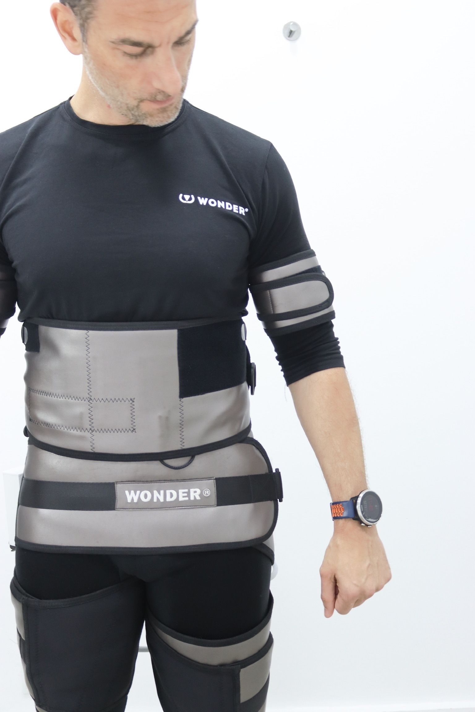 Hombre con traje deportivo gris y negro, con el brazo levantado y la mirada baja. Fondo blanco.