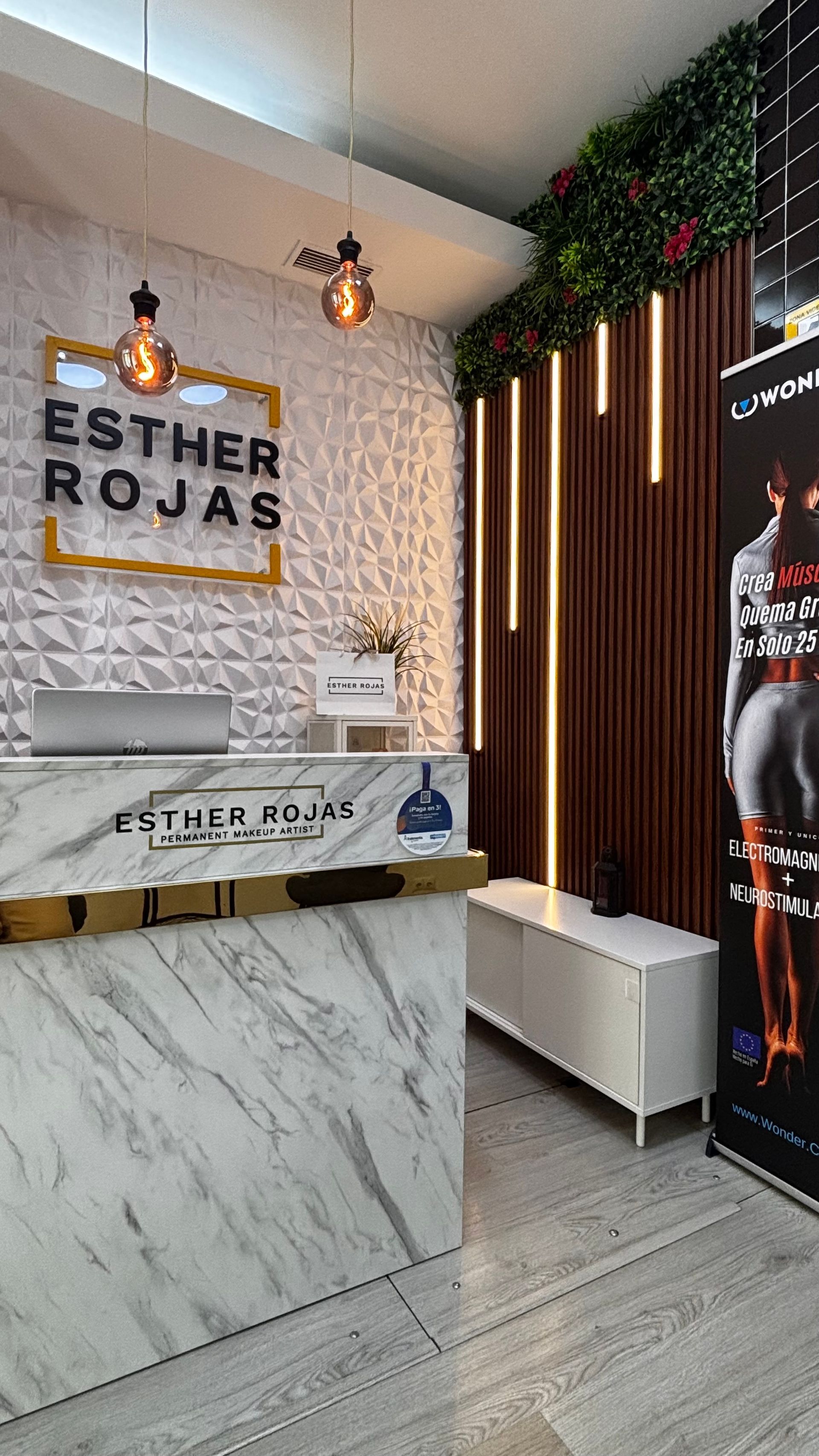 Zona de recepción del spa Esther Rojas, mostrador de mármol, cartel con nombre, paredes blancas y pancarta de una persona.