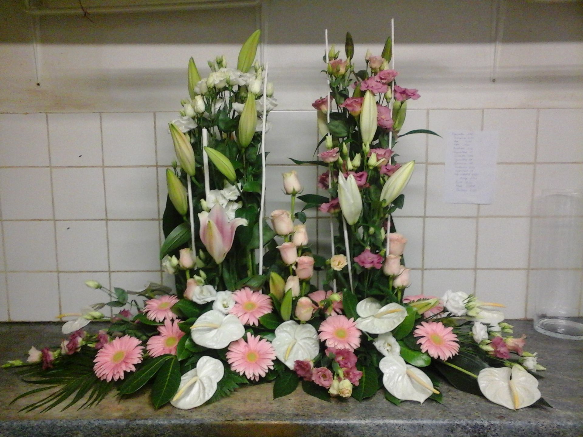 Une composition florale à plusieurs niveaux, composée de gerberas roses, de lys blancs, de roses roses et d'anthuriums blancs, posée sur un comptoir.