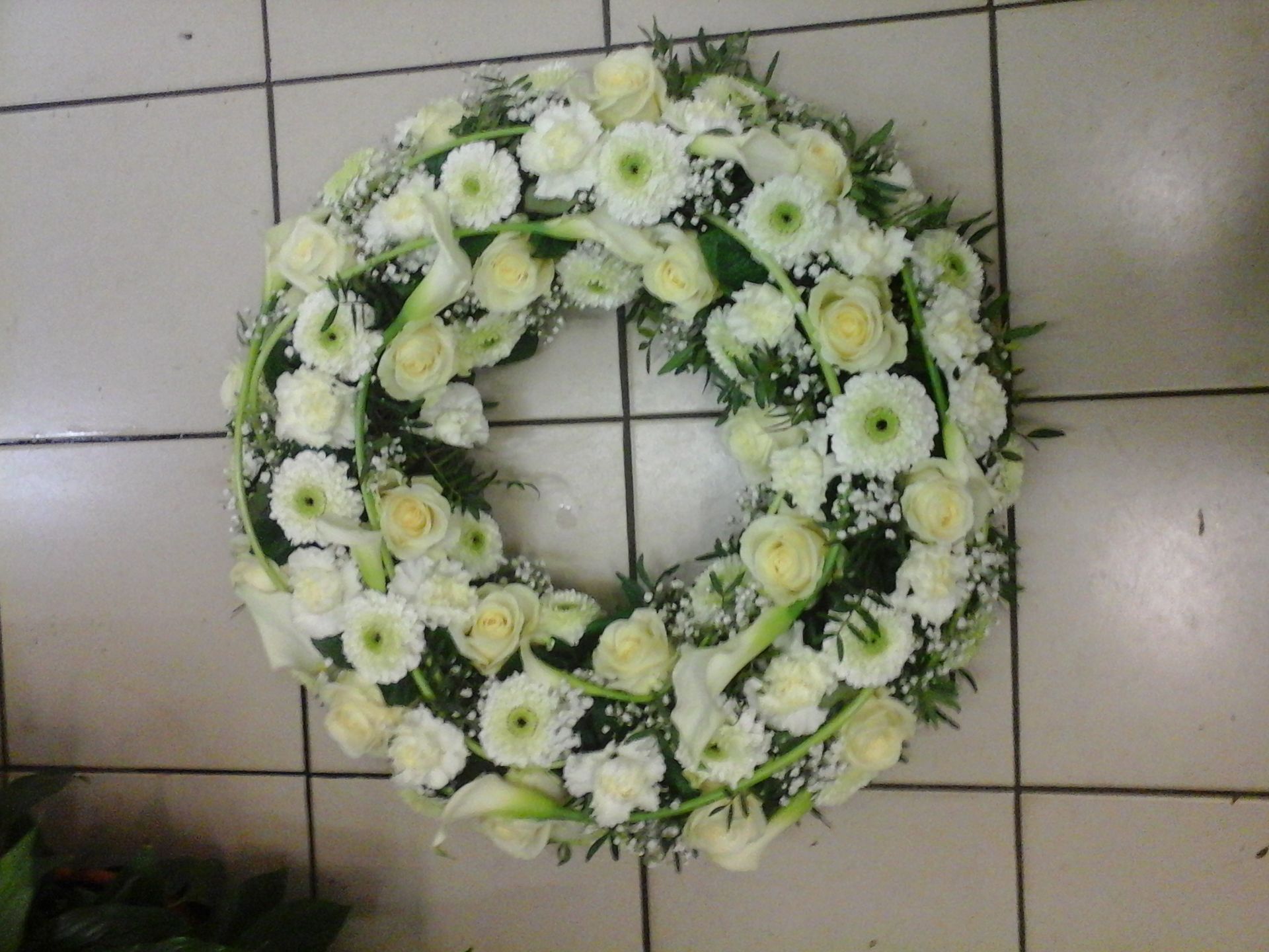 Une couronne funéraire circulaire composée de roses blanches, de gerberas et de lys calla, agrémentée de verdure, sur un sol carrelé.