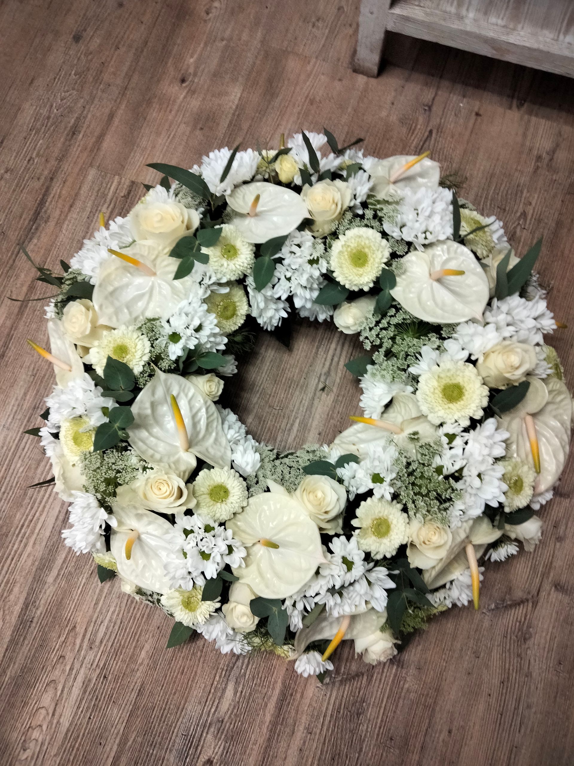 Une couronne funéraire circulaire composée d'anthuriums blancs, de roses et de chrysanthèmes avec du feuillage vert, posée sur un sol en bois.