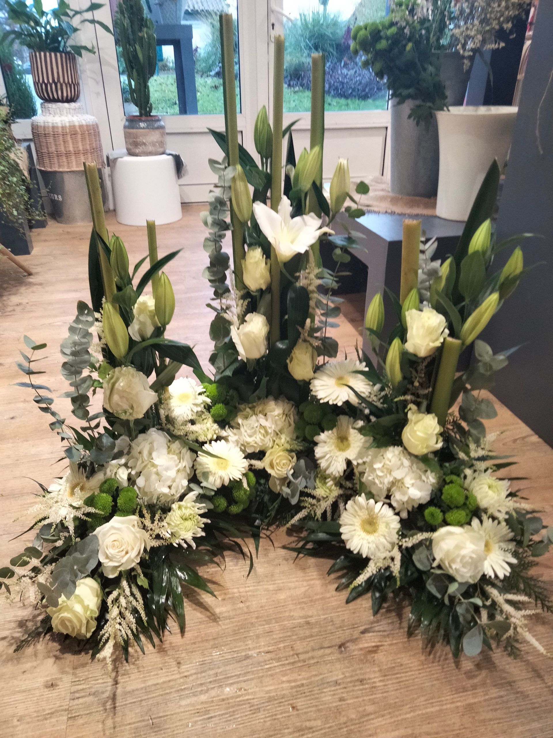 Trois élégantes compositions florales avec des roses blanches, des lys et de la verdure, disposées sur un parquet dans une boutique de fleuriste.