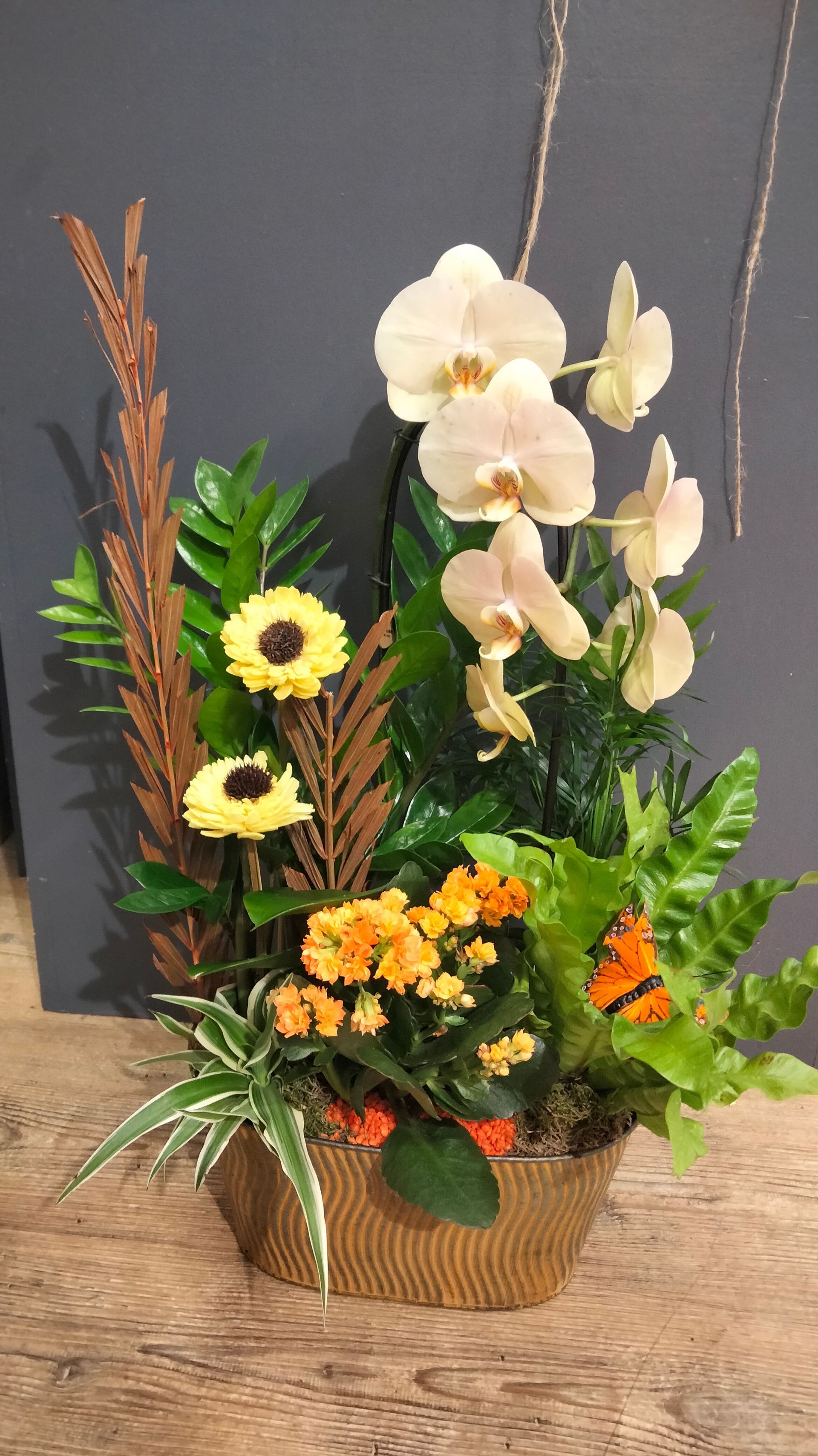 Une composition florale en panier comprenant des orchidées blanches, des tournesols jaunes, des fleurs orange et un assortiment de feuillages verts.