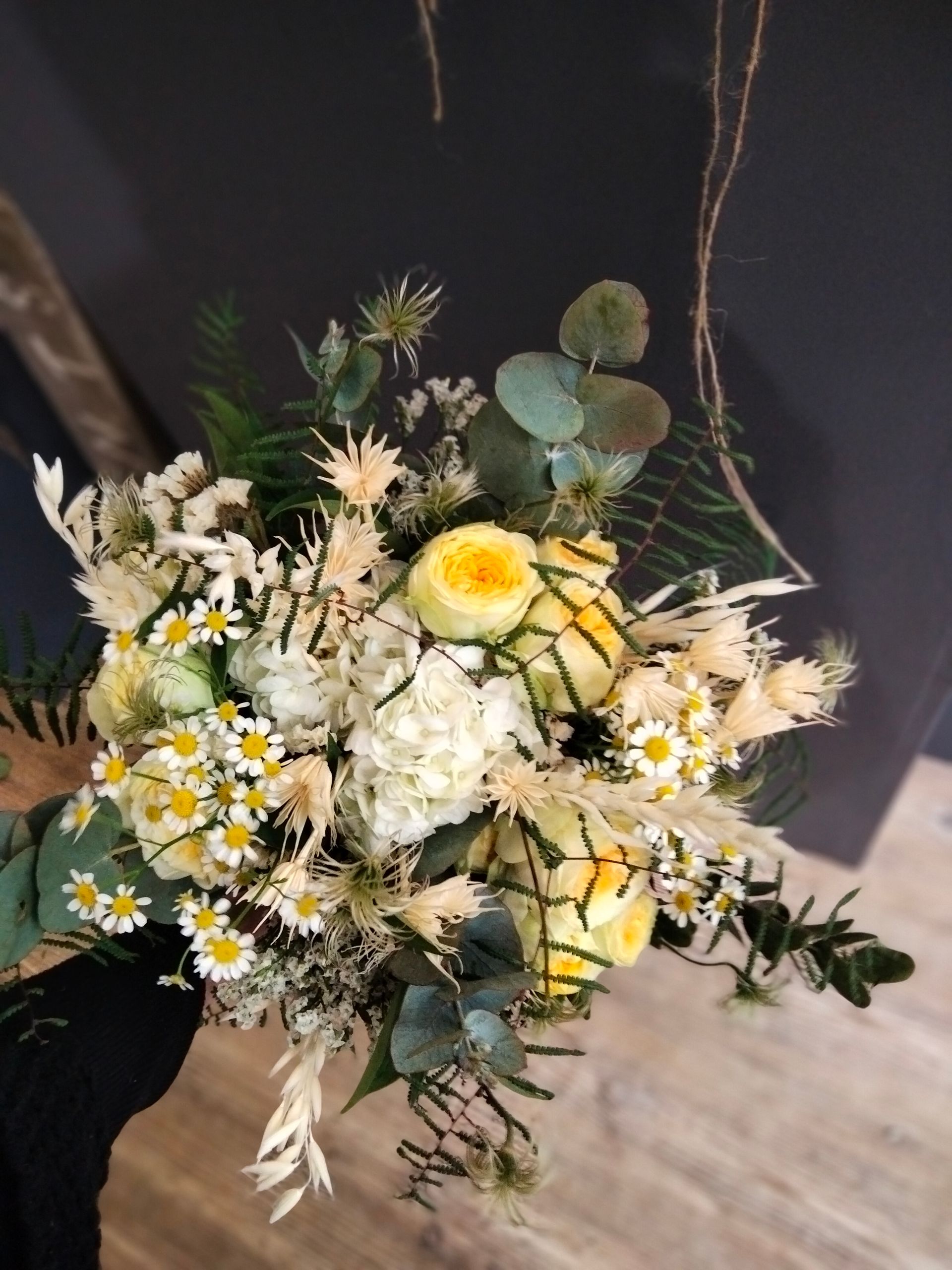Un bouquet de roses jaunes, de marguerites blanches et de fleurs séchées, agrémenté de feuillage d'eucalyptus, sur un fond sombre.