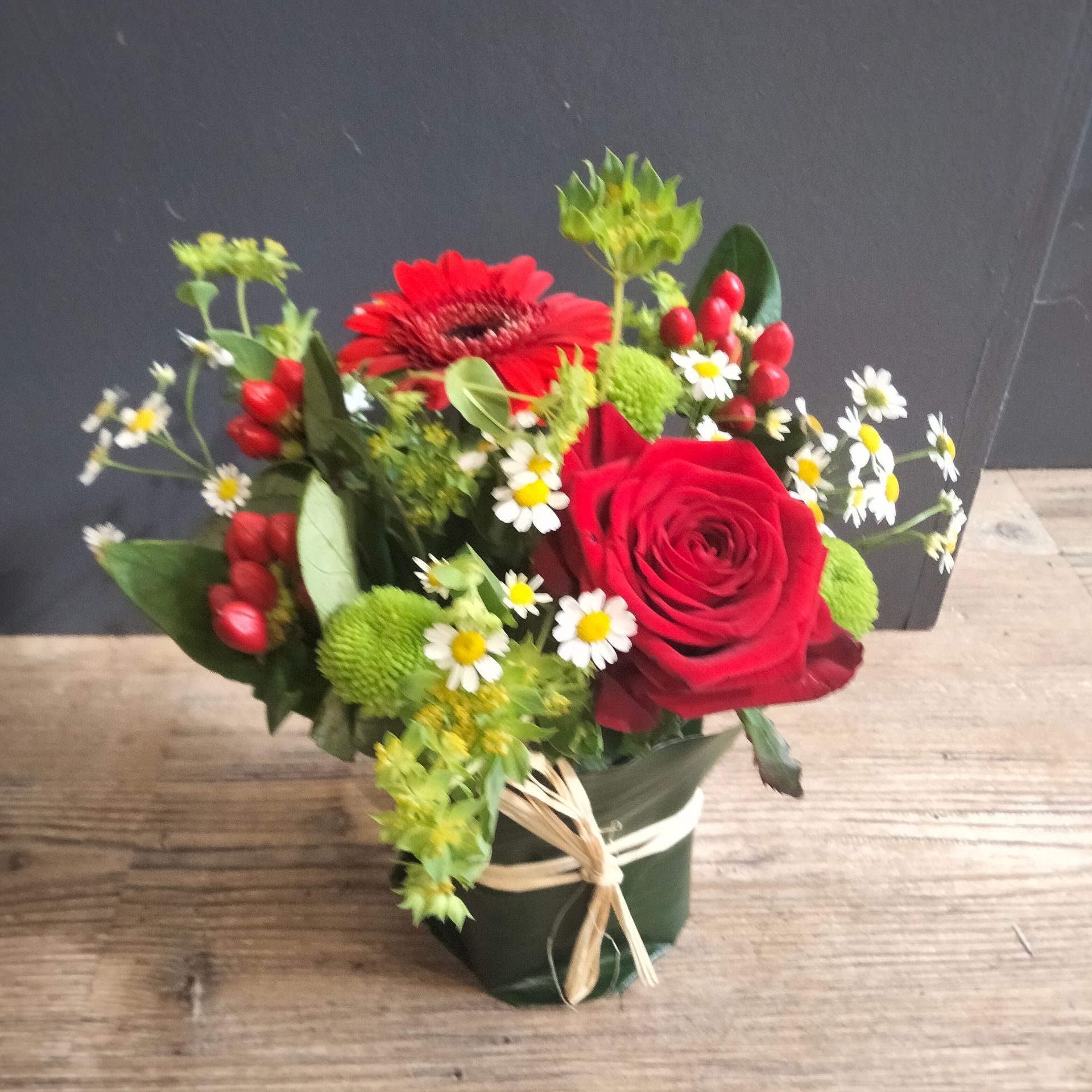 Un petit arrangement floral composé d'une rose rouge, d'une gerbera rouge, de marguerites blanches et de baies rouges dans un pot recouvert de feuilles.