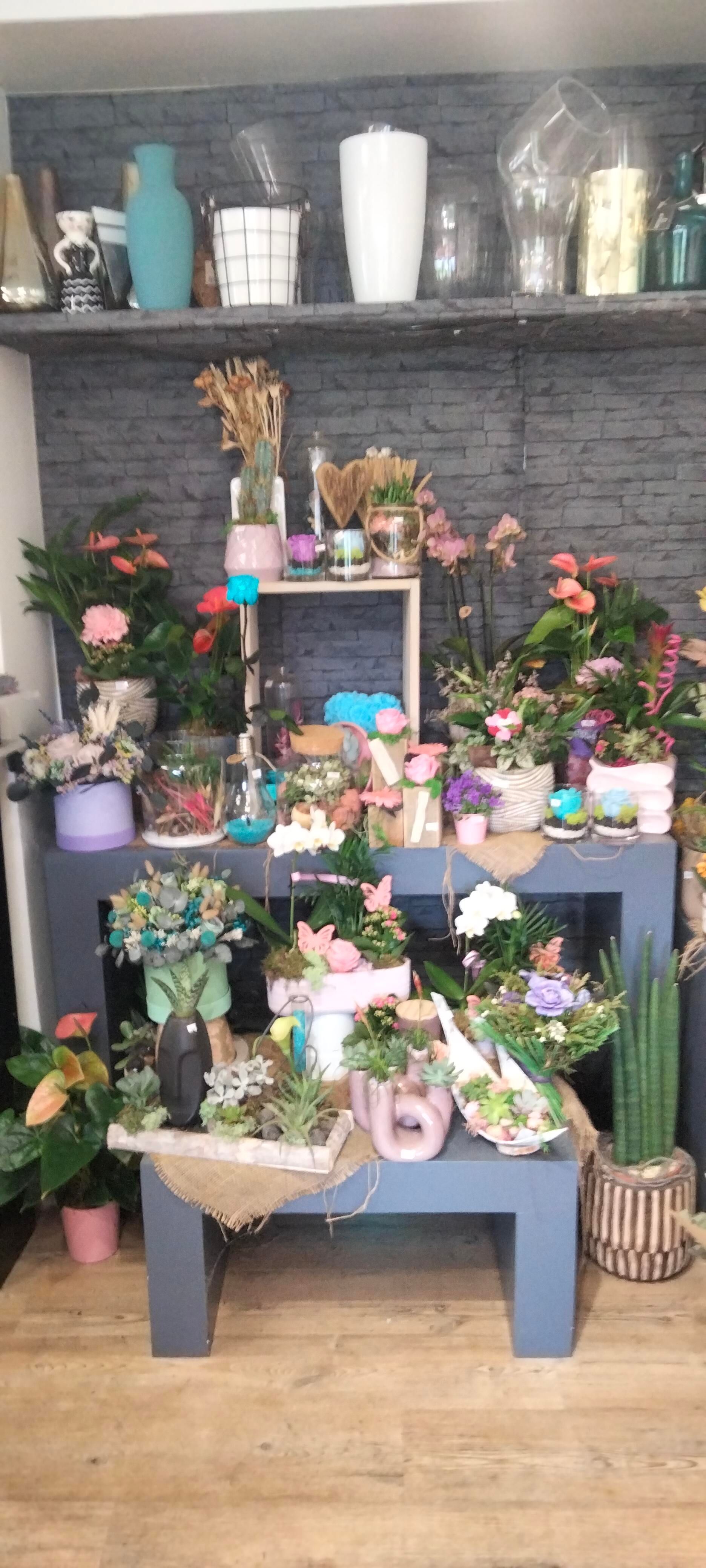 Une étagère de vente au détail présentant diverses plantes en pot, des fleurs et des vases en céramique colorés, devant un mur de briques grises.