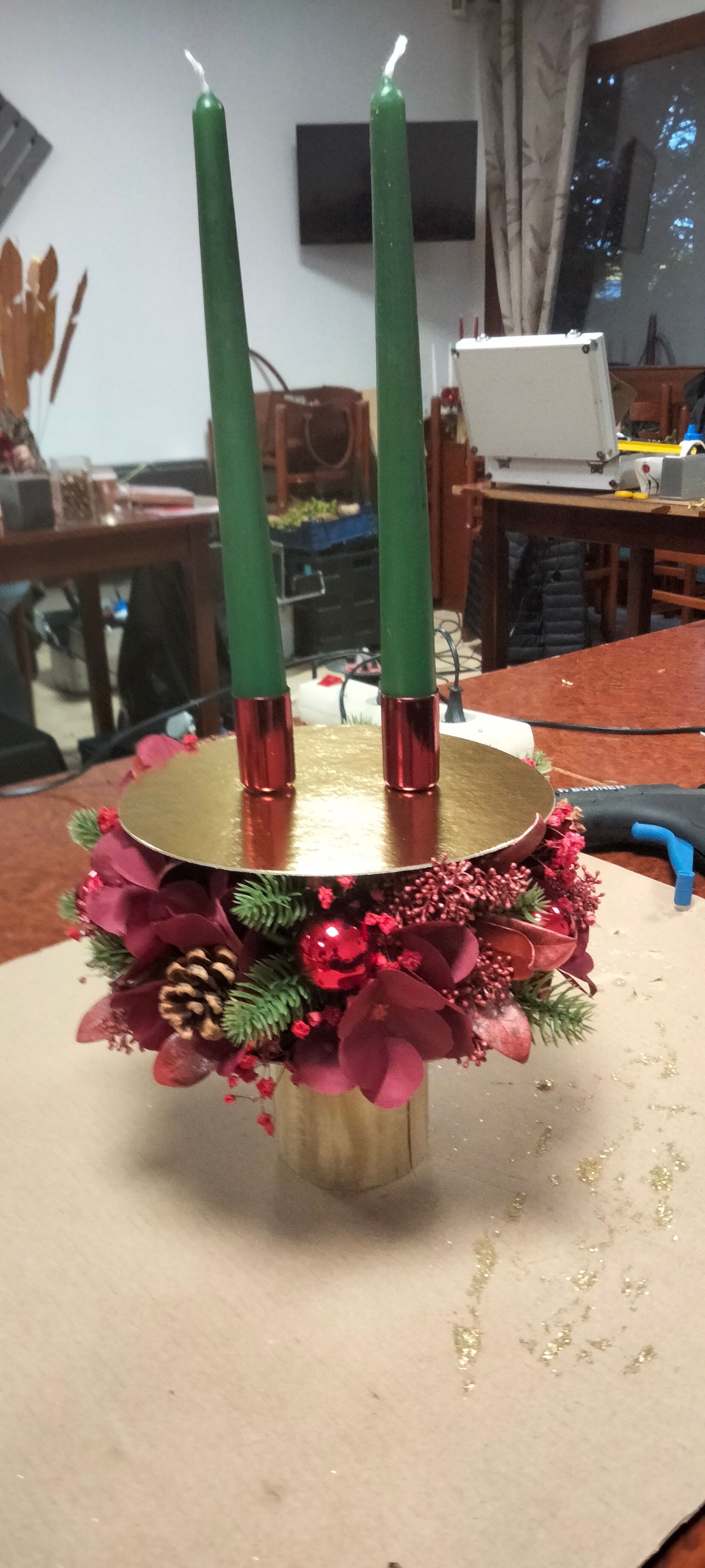 Deux bougies vertes effilées sont posées sur un centre de table doré orné de fleurs rouges, de baies et de pommes de pin.