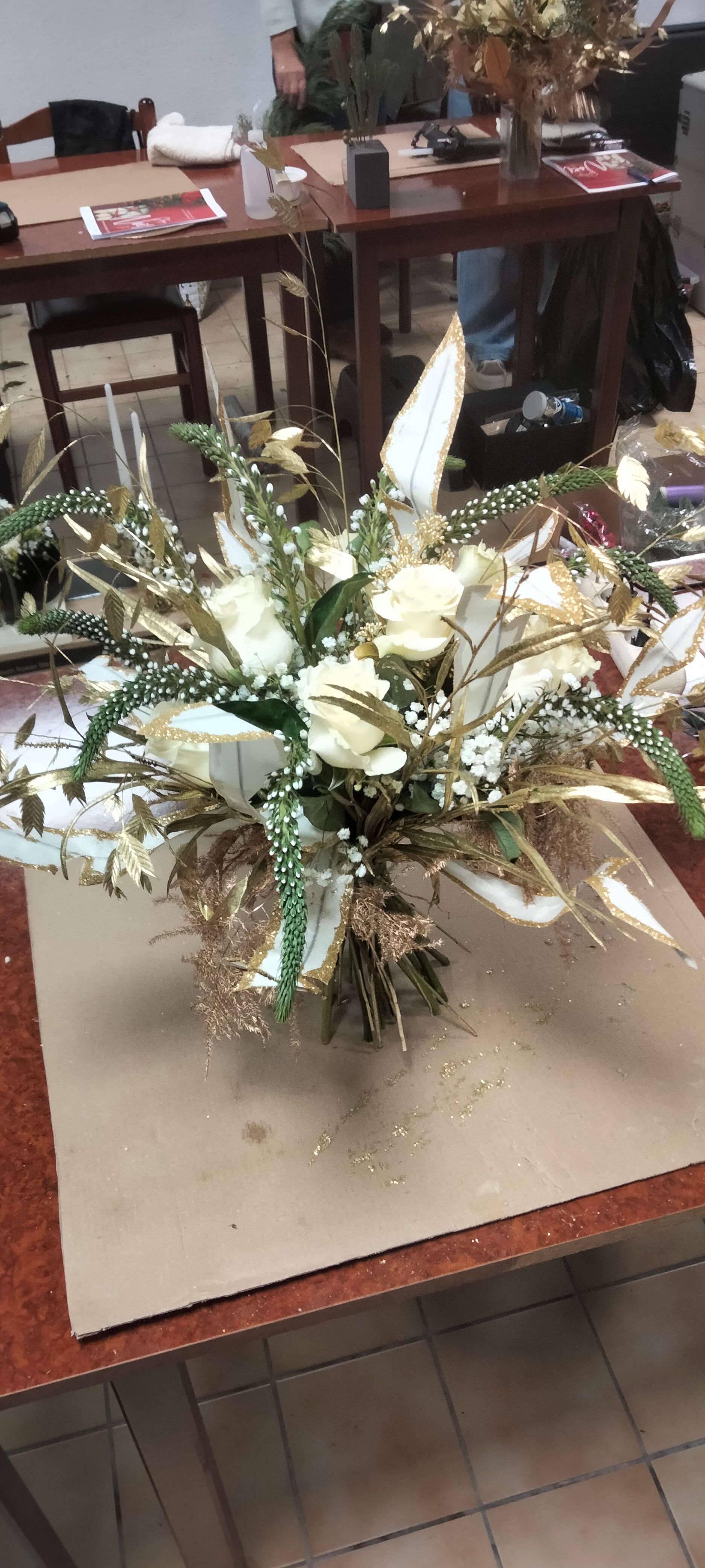 Une composition florale festive avec des roses blanches, de la verdure et des touches dorées métallisées est posée sur une table dans un atelier.