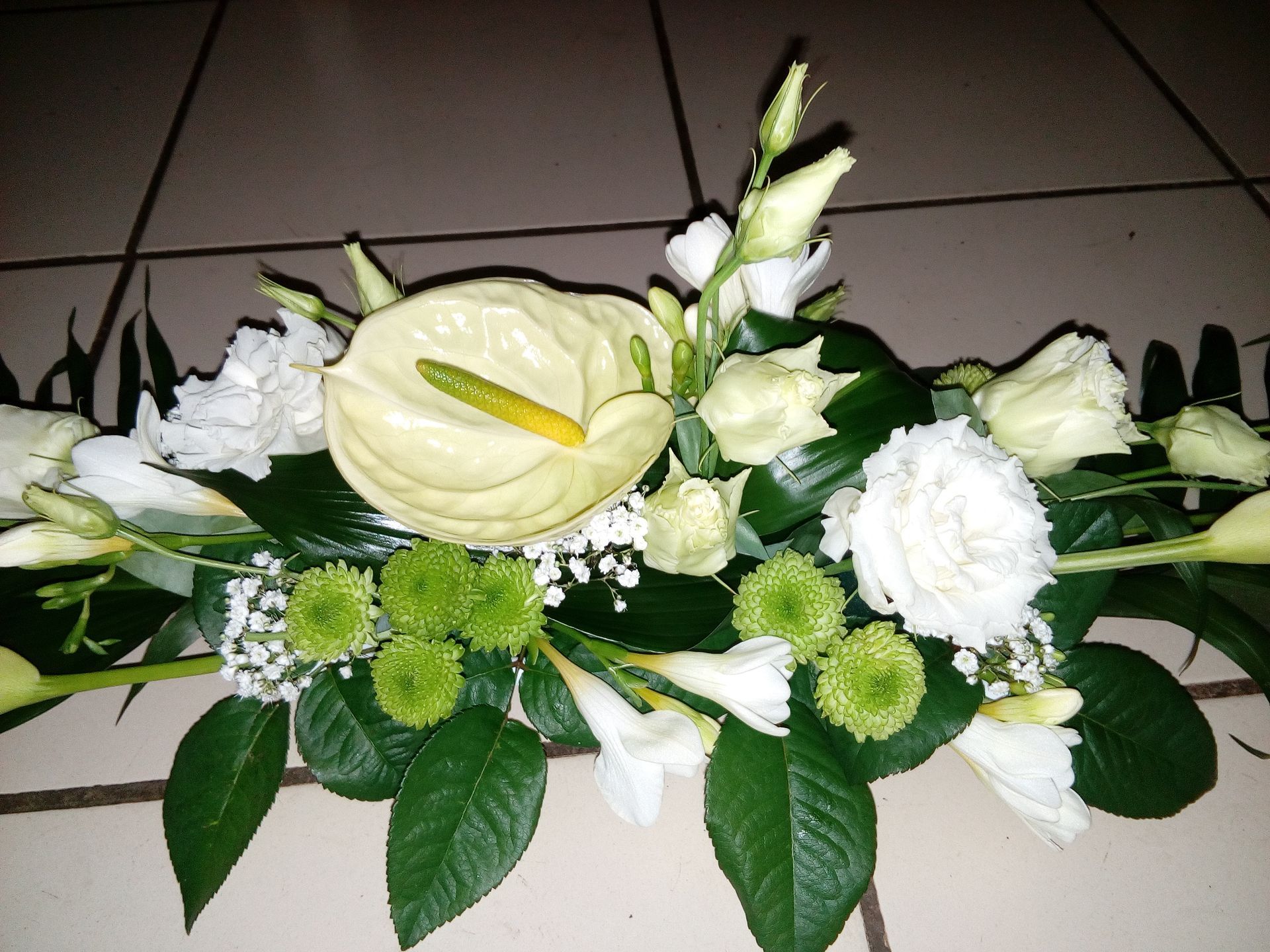 Une composition florale comprenant un anthurium blanc, des roses blanches, des chrysanthèmes verts et du gypsophile, sur un sol carrelé.