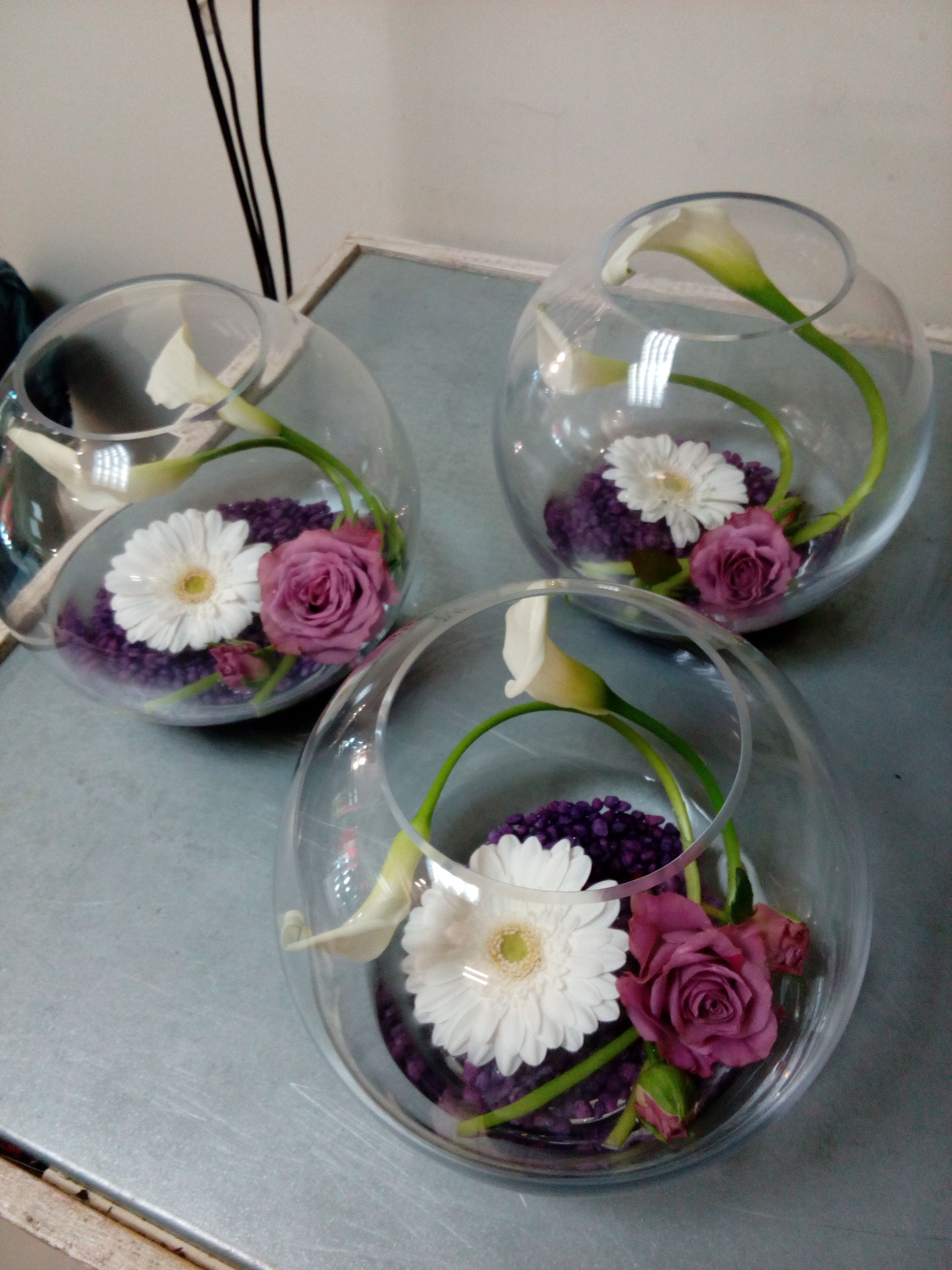 Trois vases en verre en forme de bocal à poissons, contenant chacun une gerbera blanche, une rose mauve et un arum sur des galets violets.