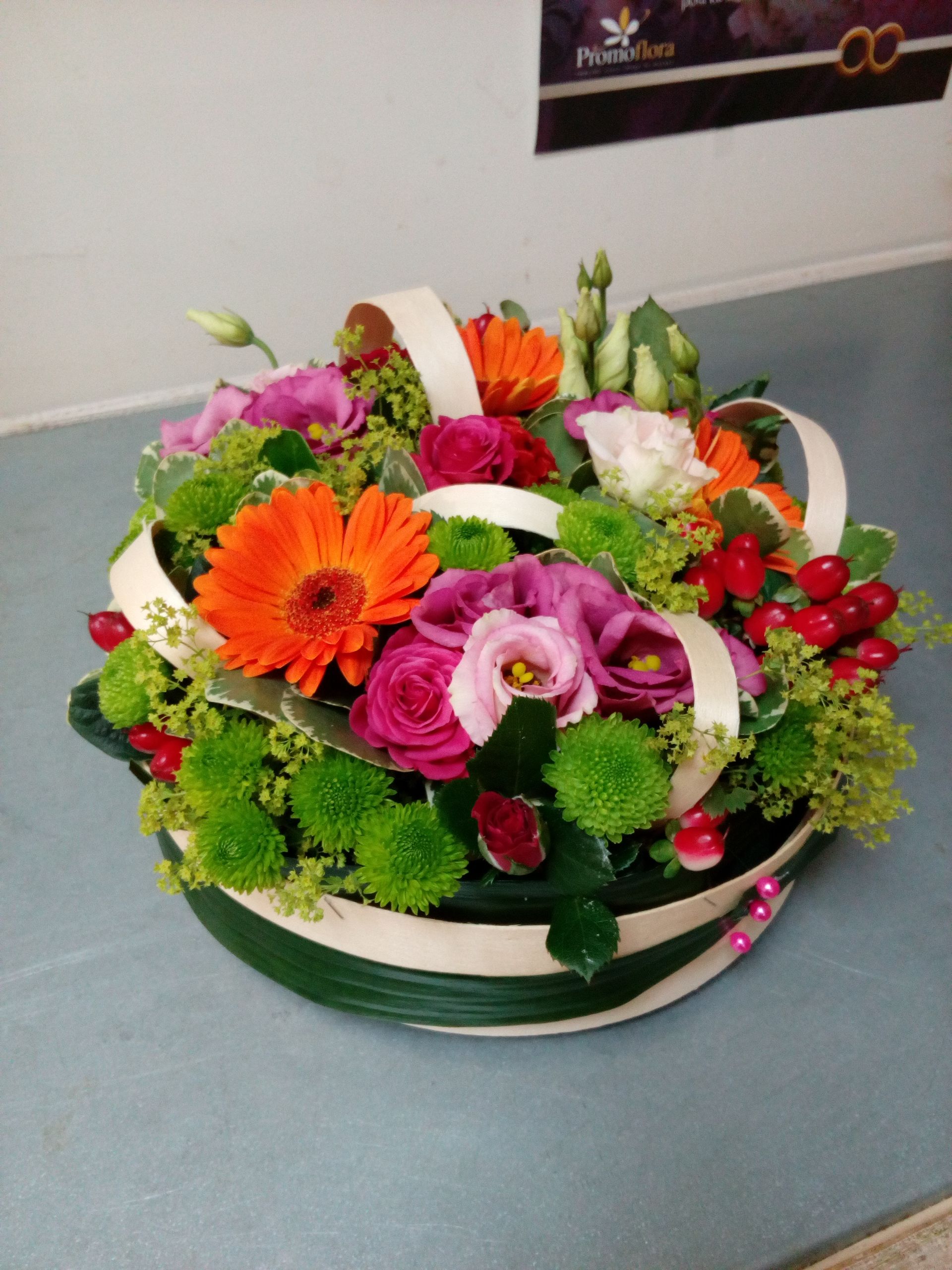 Un arrangement floral éclatant dans un récipient rond, composé de gerberas orange, de roses roses, de chrysanthèmes verts et de baies.