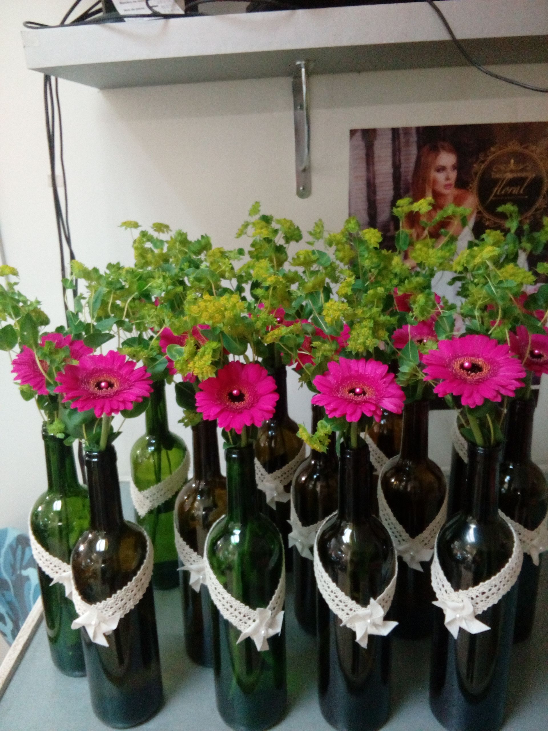 Plusieurs bouteilles en verre vert foncé, ornées d'un ruban de dentelle blanche, contenant chacune une gerbera rose vif et de la verdure.