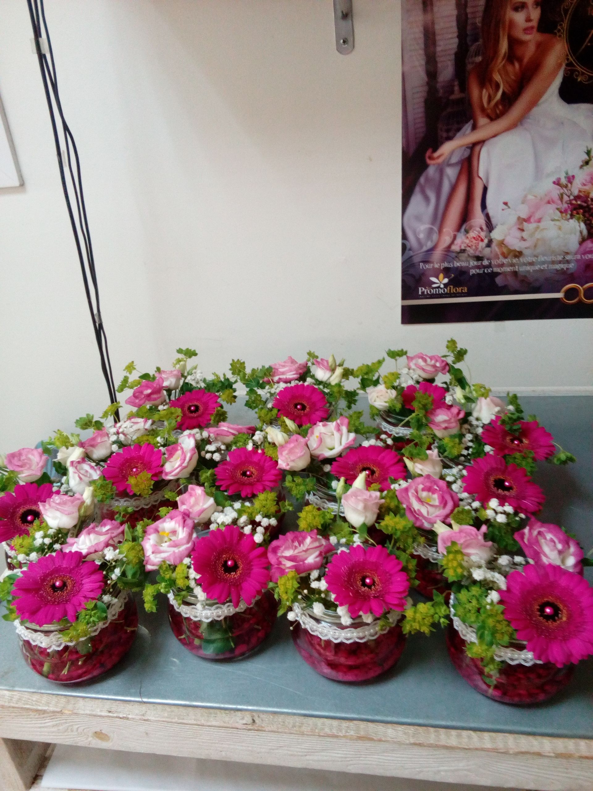 Quatre bocaux en verre posés sur une table, chacun rempli de gerberas rose vif, de roses rose pâle et de petites branches vertes.