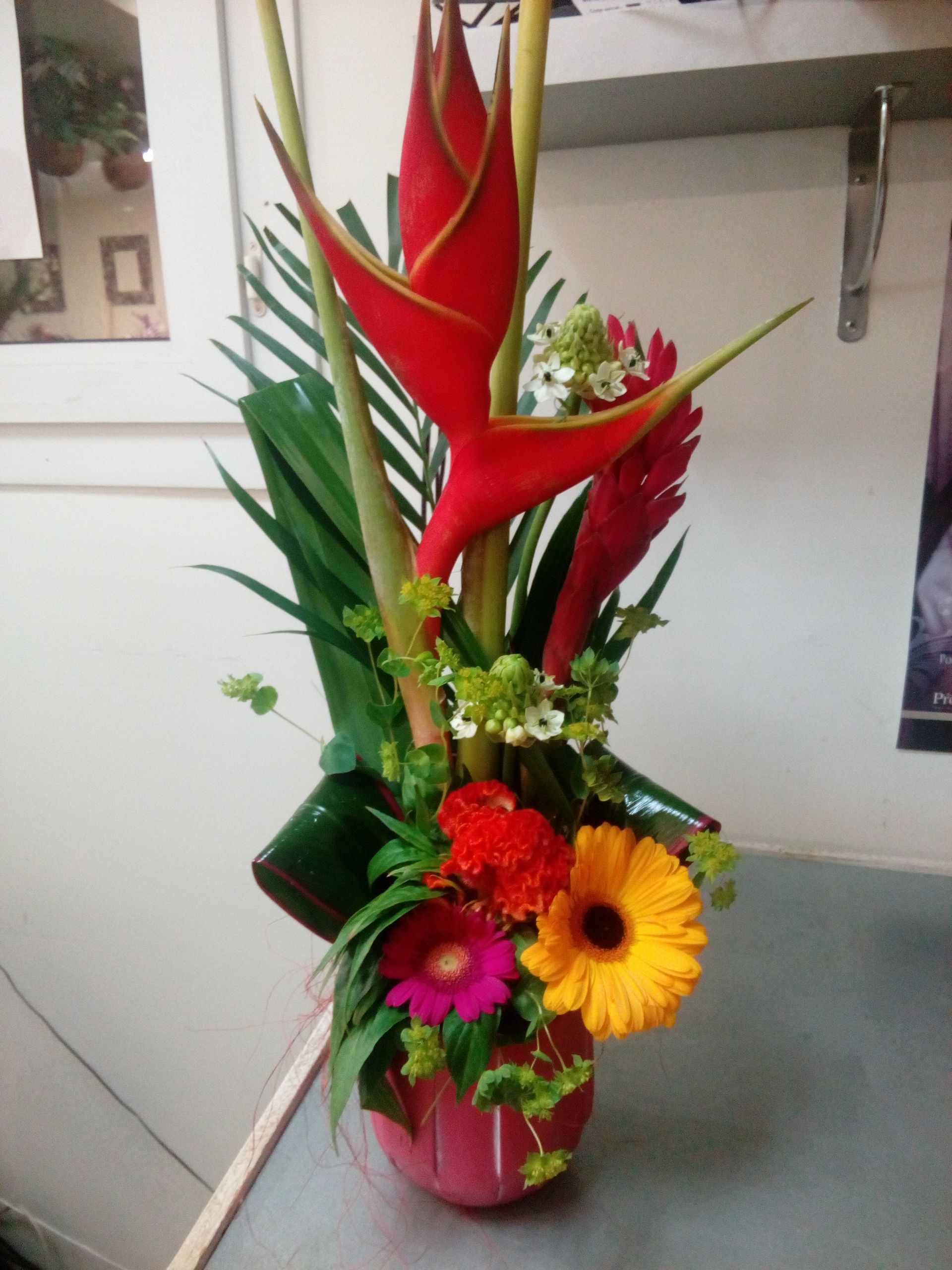Une composition florale verticale mettant en scène des héliconias rouge vif, du gingembre, des gerberas jaunes et roses dans un vase rose.