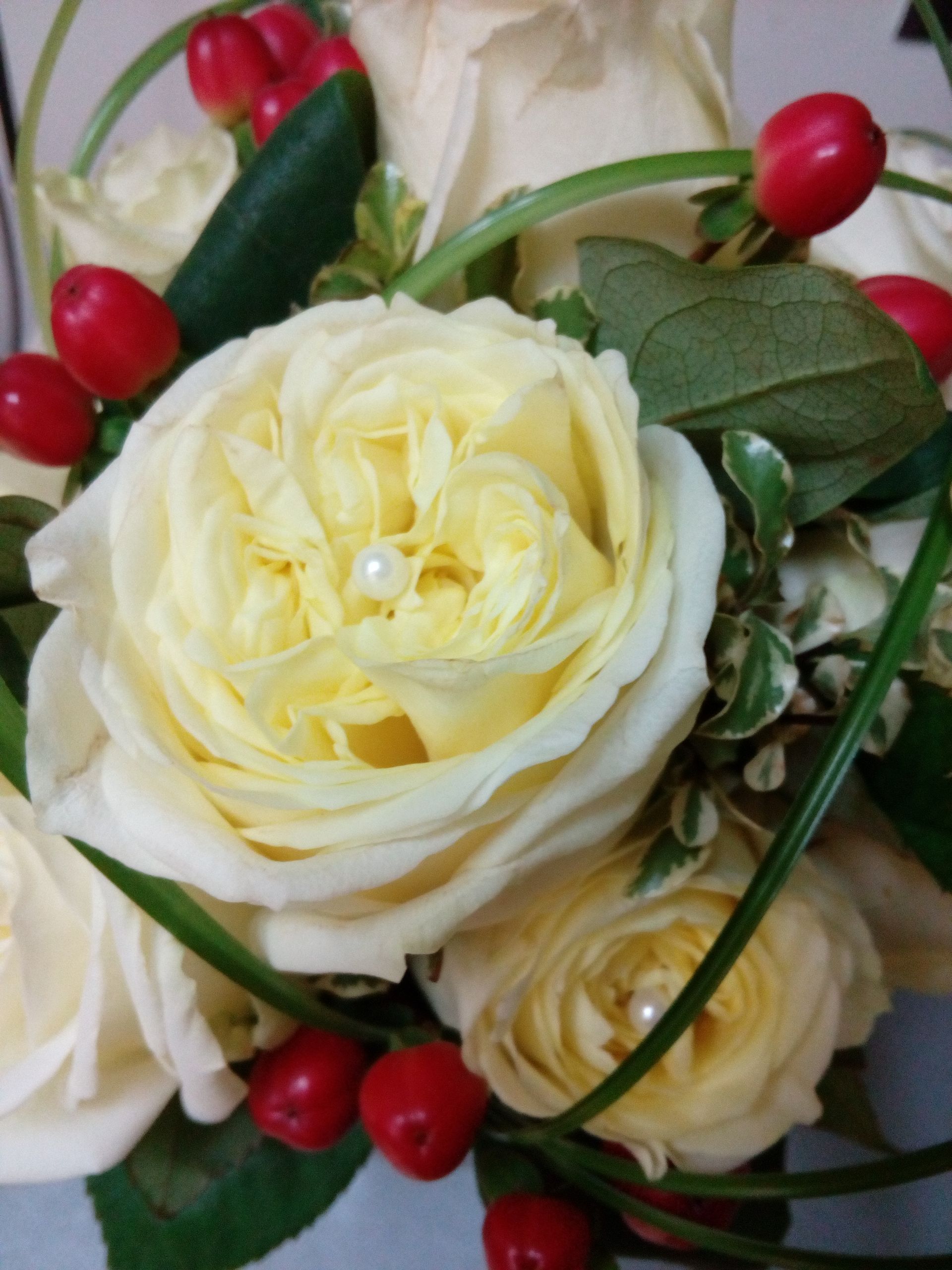 Gros plan sur un bouquet de roses jaune pâle à cœur nacré, agrémenté de baies rouge vif et de feuillage vert.