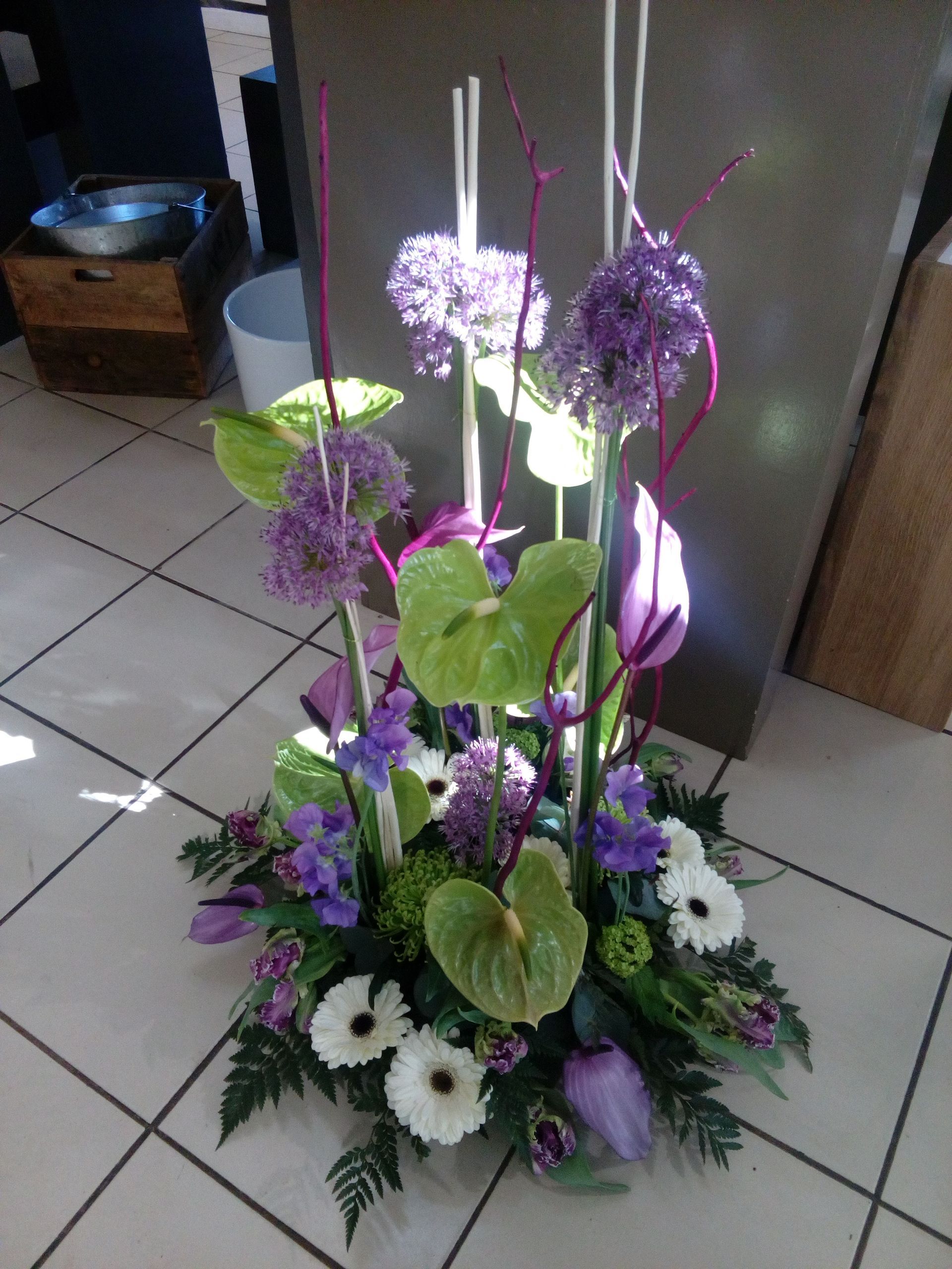 Une composition florale décorative composée d'alliums violets, d'anthuriums verts, de gerberas blancs et de verdure, disposée au sol.
