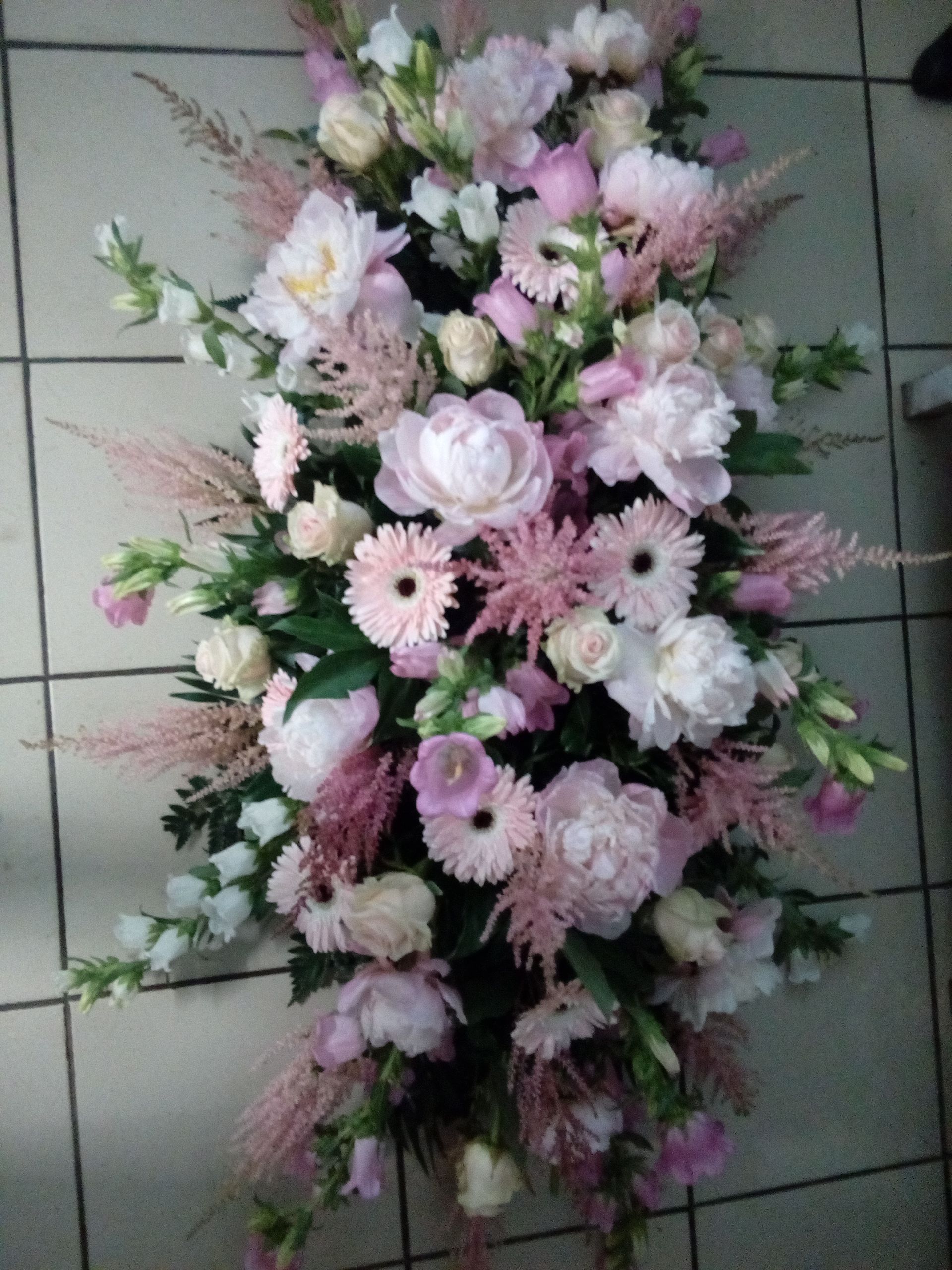 Une élégante composition florale funéraire mettant en scène des pivoines, des gerberas et des astilbes roses sur un sol carrelé.
