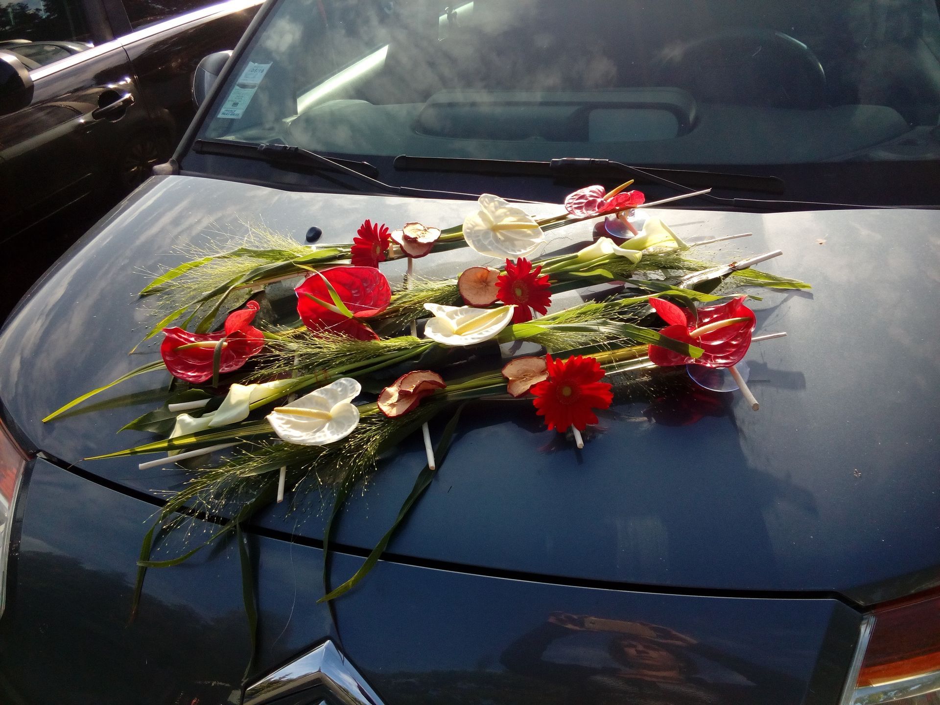 Décoration florale composée d'anthuriums rouges et de lys blancs disposés sur le capot bleu foncé d'une voiture.