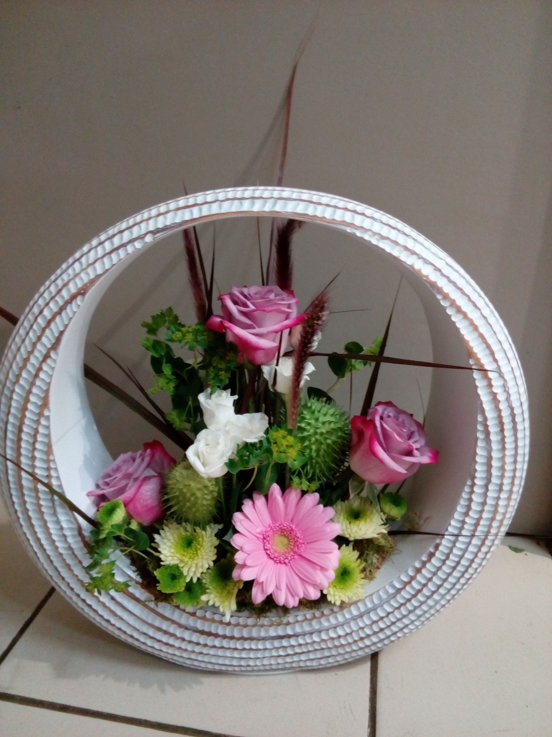 Un arrangement floral de roses roses, de fleurs blanches et de touches de vert, à l'intérieur d'un cadre blanc circulaire texturé.