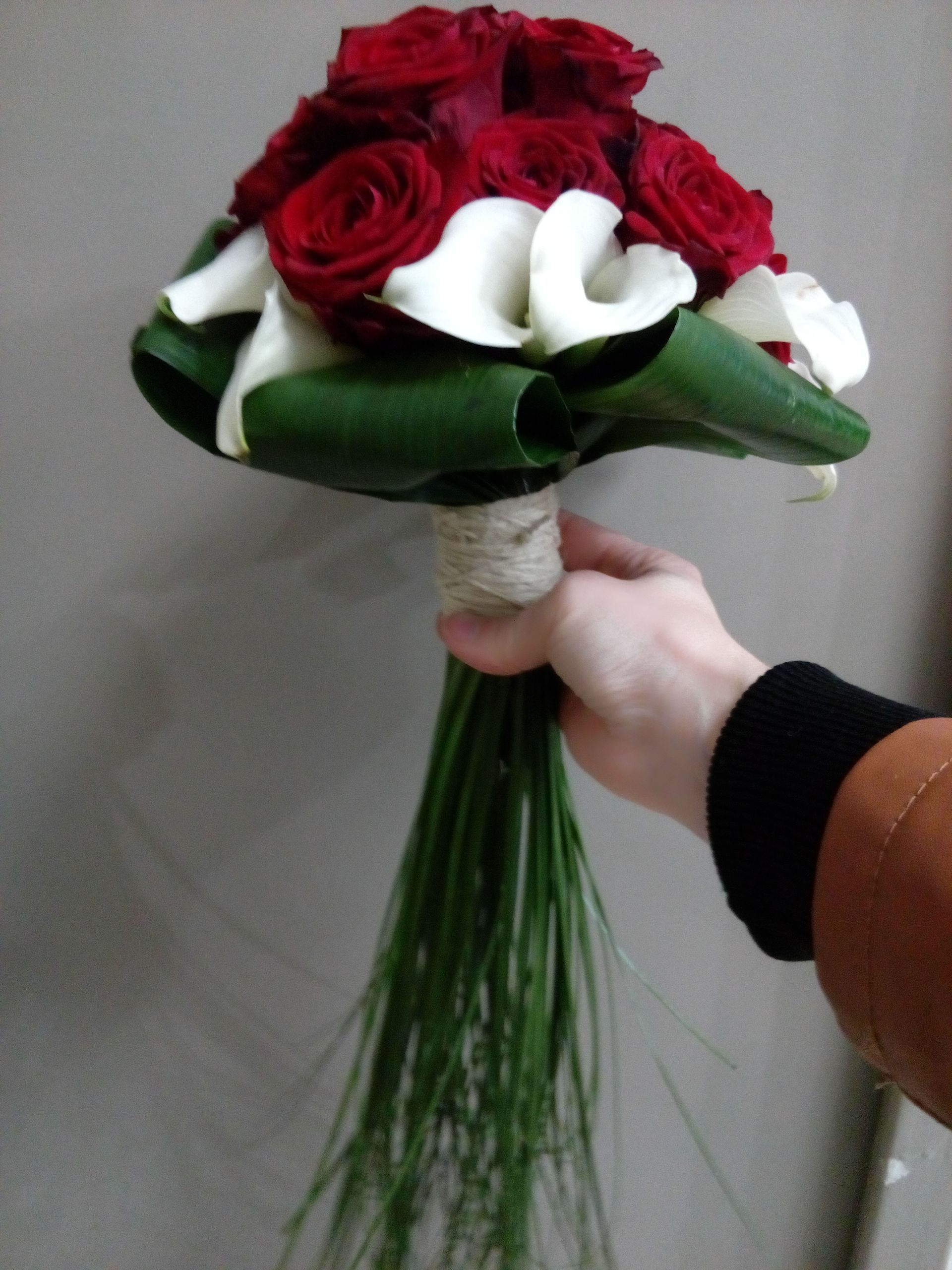 Une main tient un bouquet de roses rouge foncé et de callas blancs, enveloppé de ficelle et agrémenté de longues herbes vertes.
