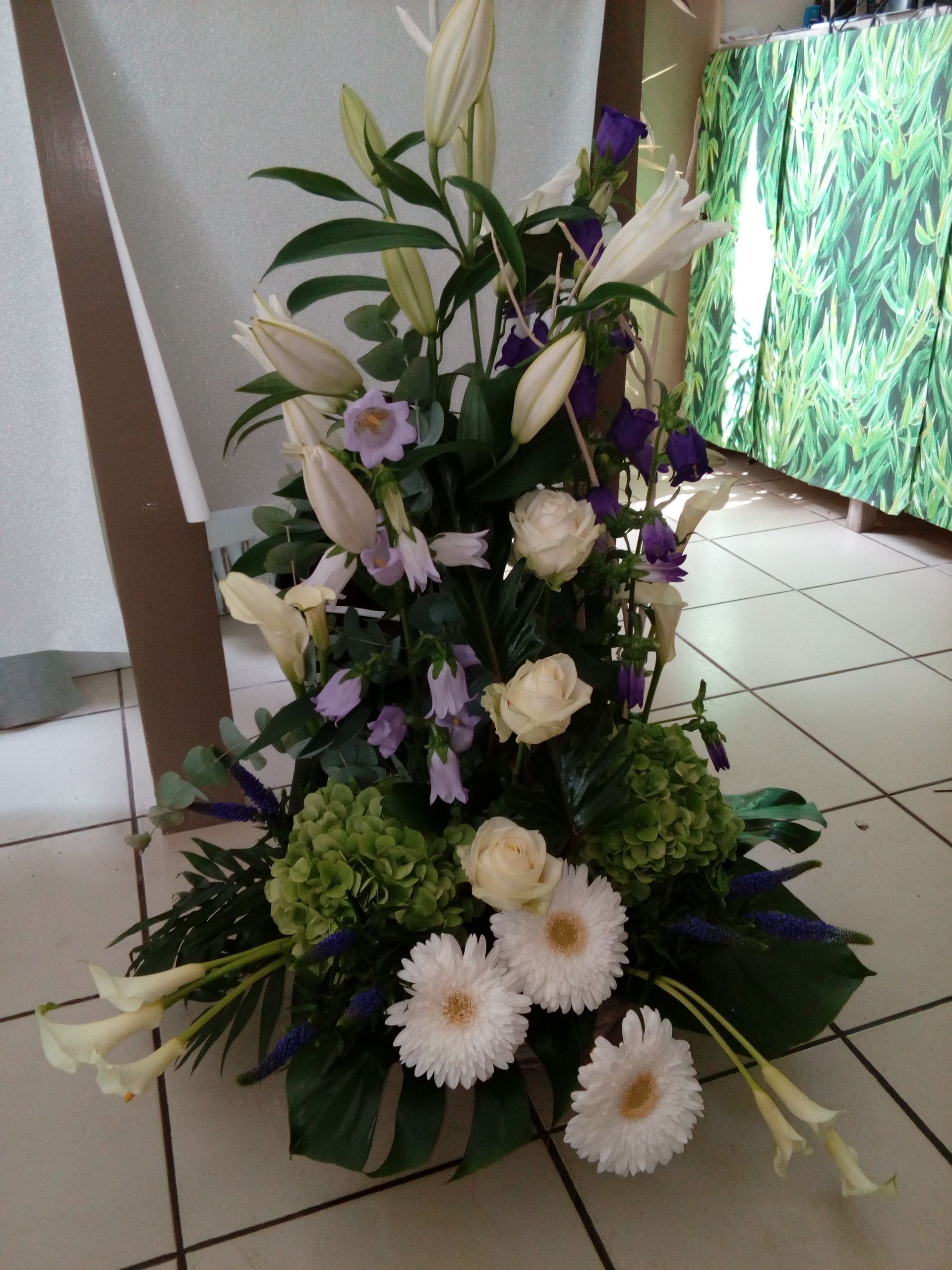 Une composition florale haute et verticale avec des lys blancs, des roses, des gerberas, des hortensias verts et des campanules violettes.