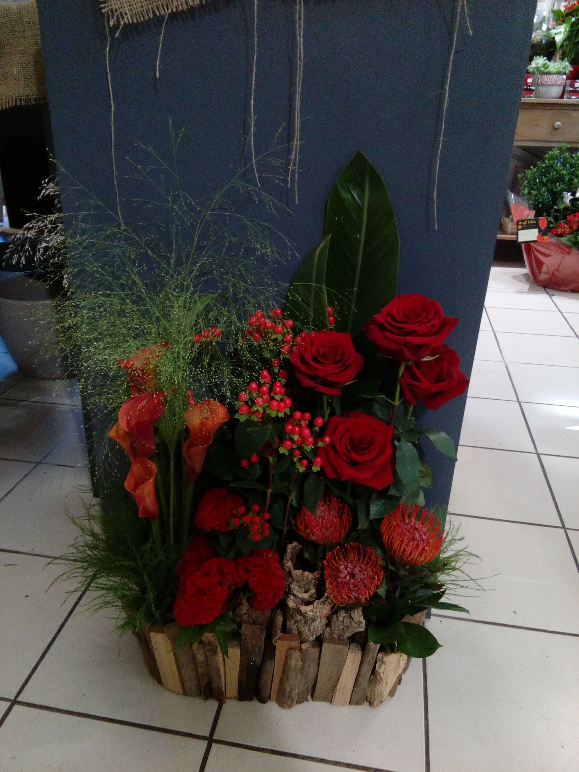 Un arrangement floral composé de roses rouges, de callas orange et de verdure dans un pot en bois tapissé d'écorce.