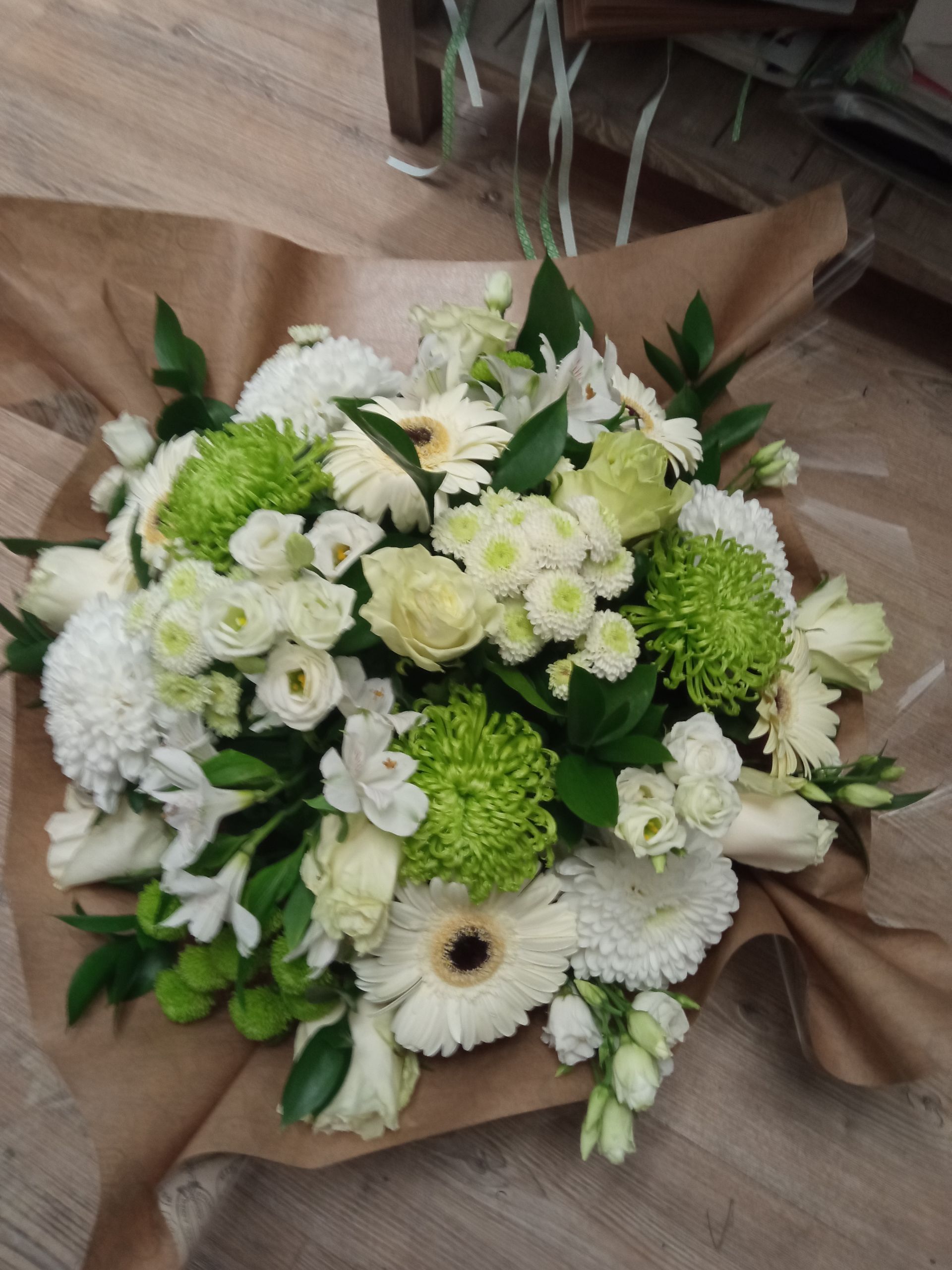 Un bouquet de fleurs blanches et vert citron, enveloppé dans du papier kraft brun, posé sur une surface en bois.