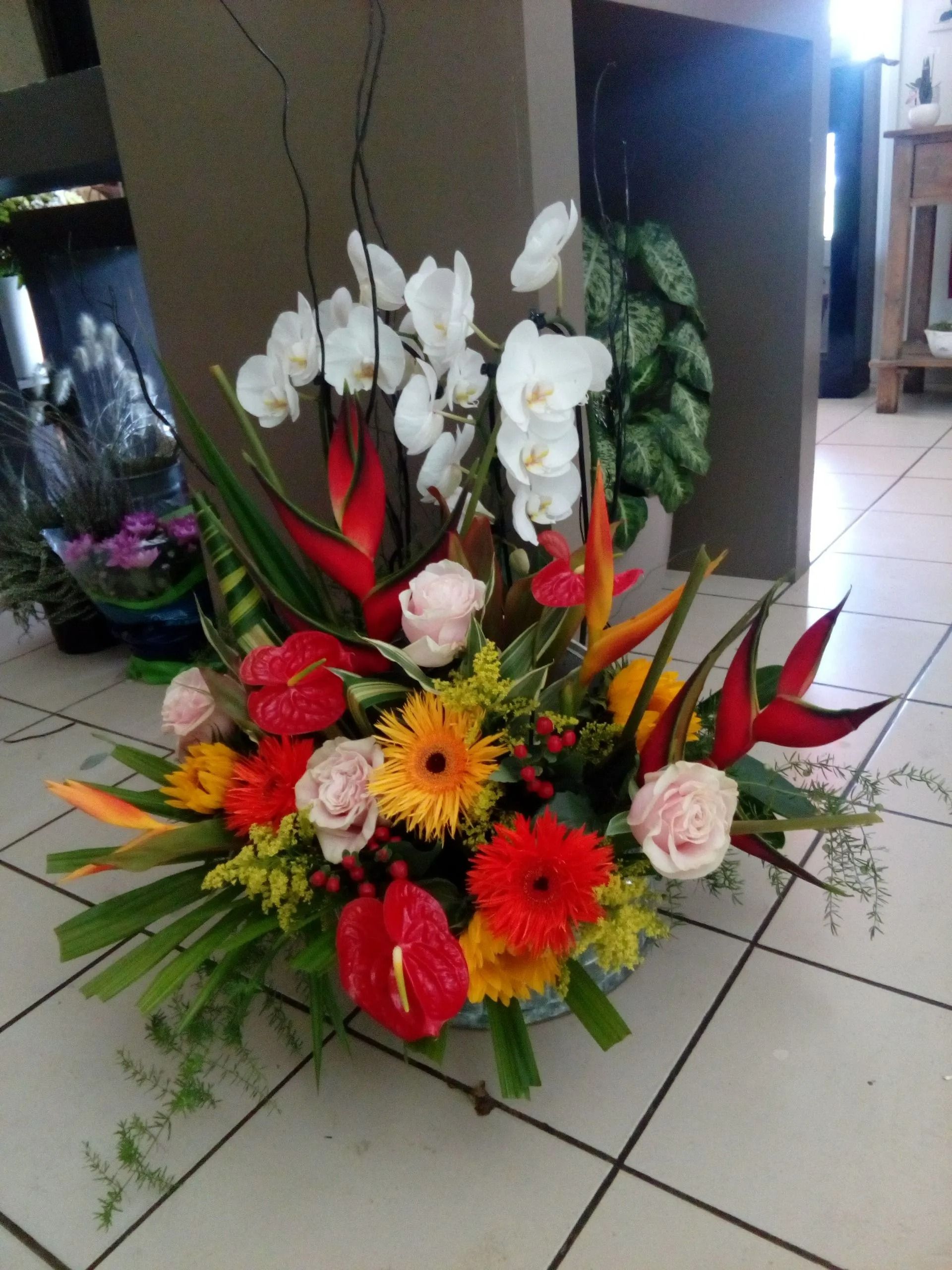 Une composition florale éclatante et multicolore, composée d'orchidées blanches, d'héliconias rouges, de roses roses et de marguerites jaunes.