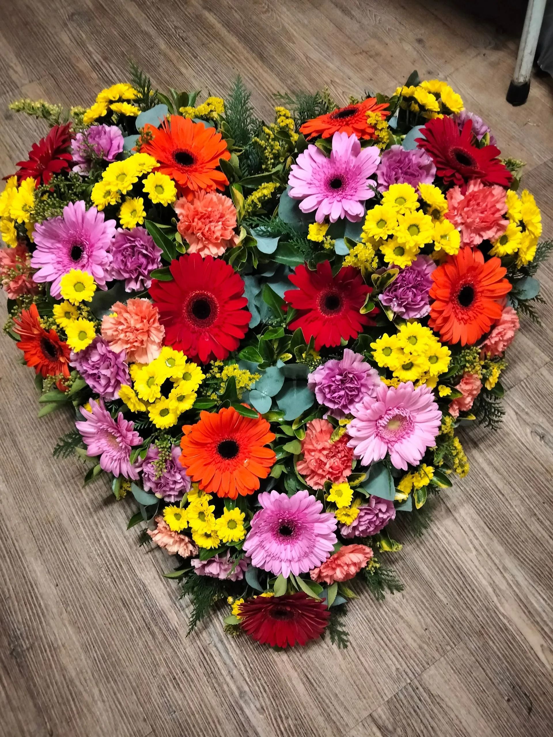Une composition florale en forme de cœur, composée de gerberas éclatants, d'œillets et de petites fleurs jaunes, sur un sol en bois.