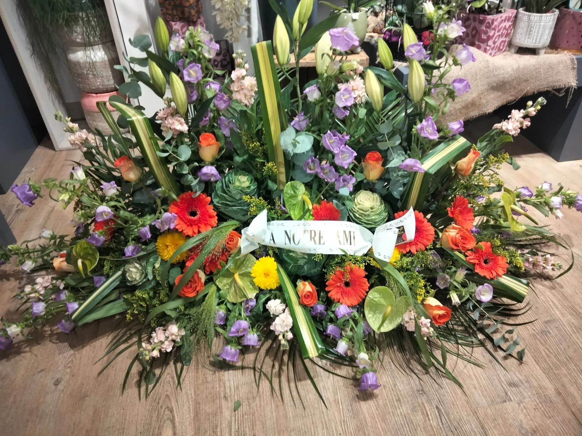 Une couronne funéraire composée de gerberas orange, de campanules violettes, de lys et de verdure, avec un ruban blanc en son centre.