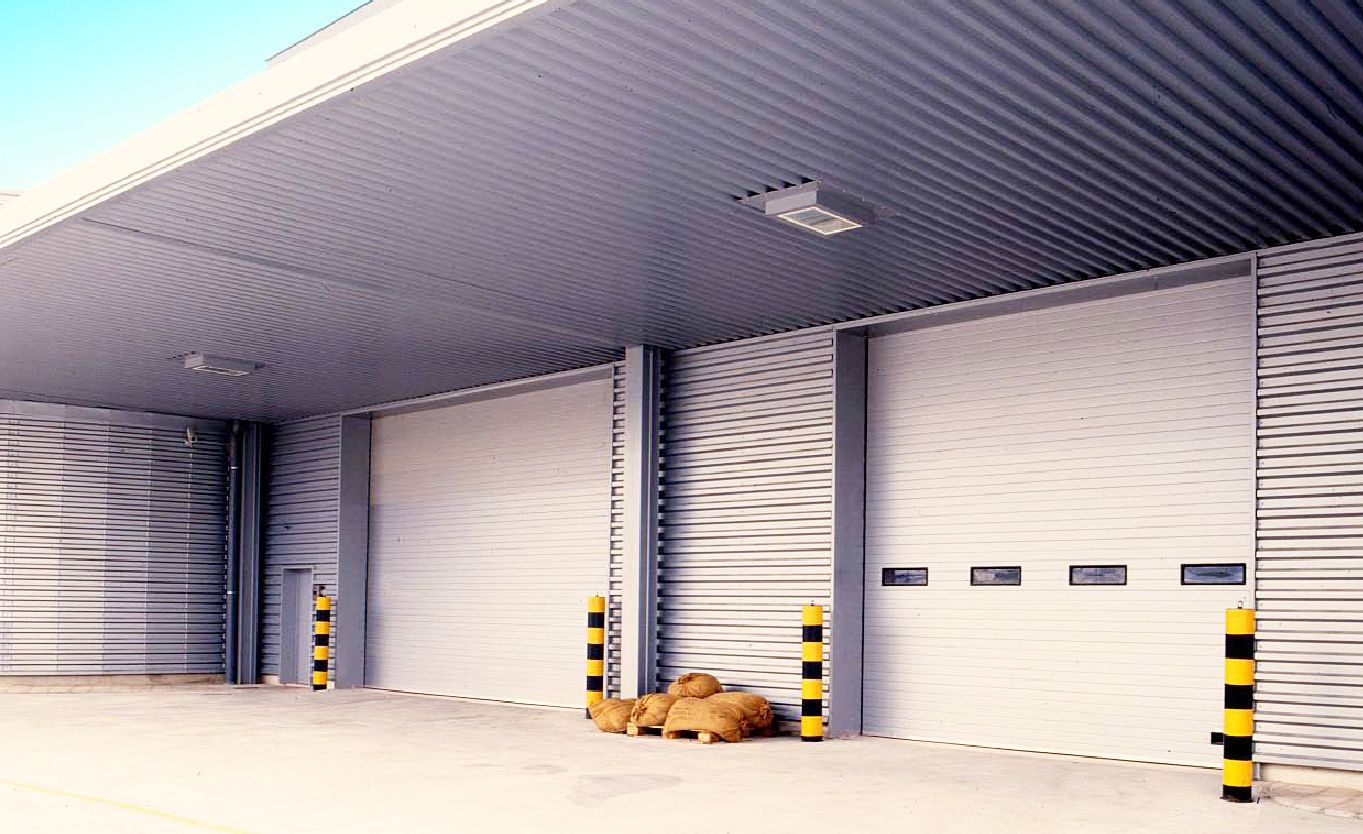 Exterior de un almacén con grandes puertas enrollables cerradas. Cerca hay bolardos amarillos y negros.