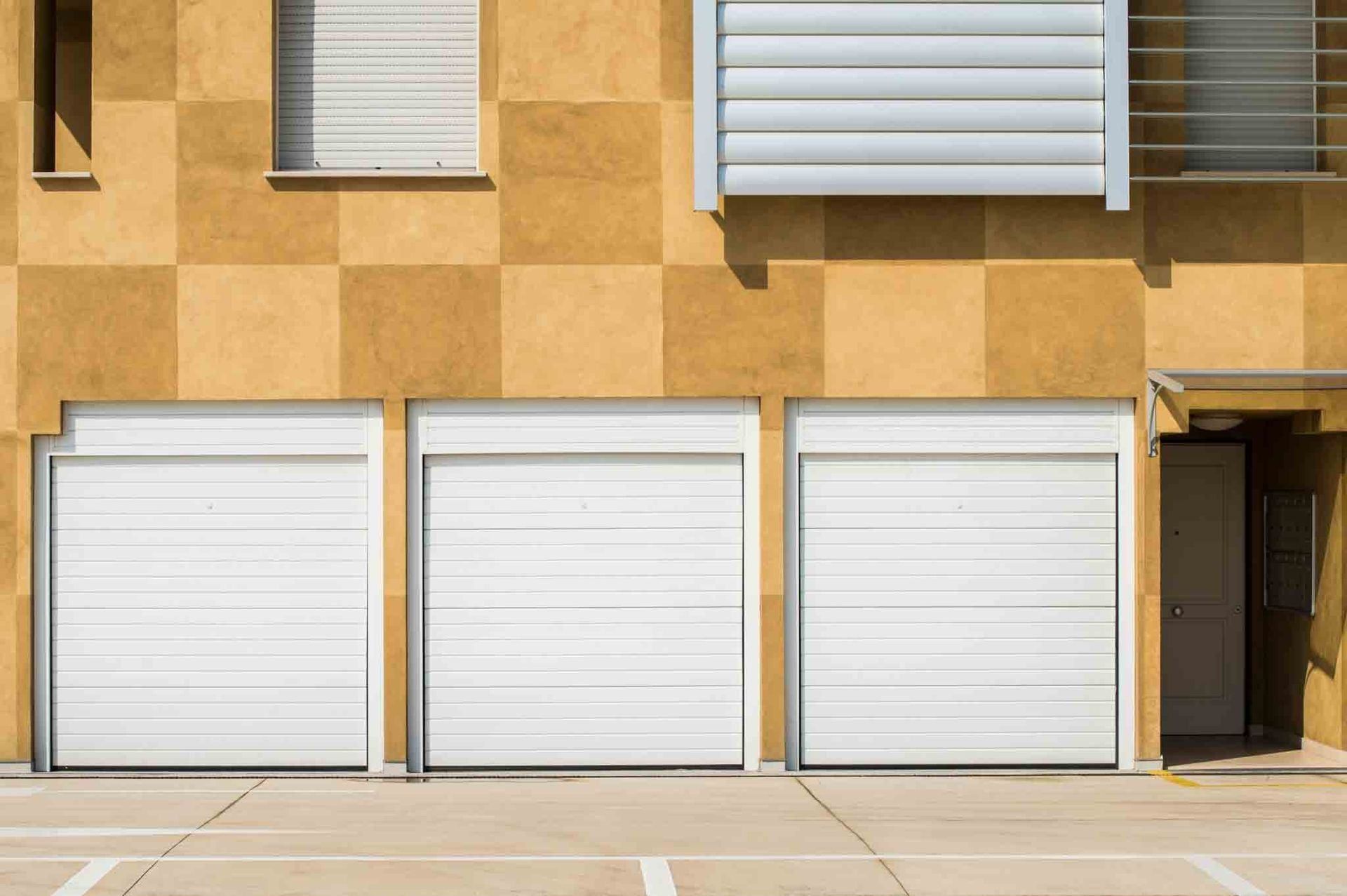 Tres puertas de garaje blancas en un edificio de cuadros amarillos. Arriba hay ventanas y un pequeño balcón.