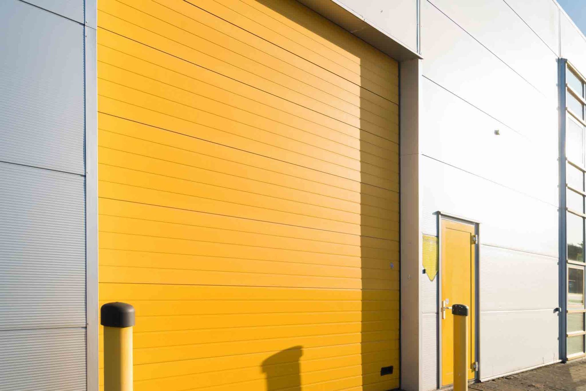 Puerta industrial amarilla y puerta amarilla adyacente en un edificio blanco con postes amarillos al frente.