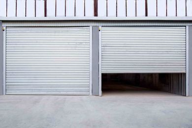 Dos puertas de garaje enrollables plateadas, una completamente cerrada y otra parcialmente abierta. Suelo de hormigón.