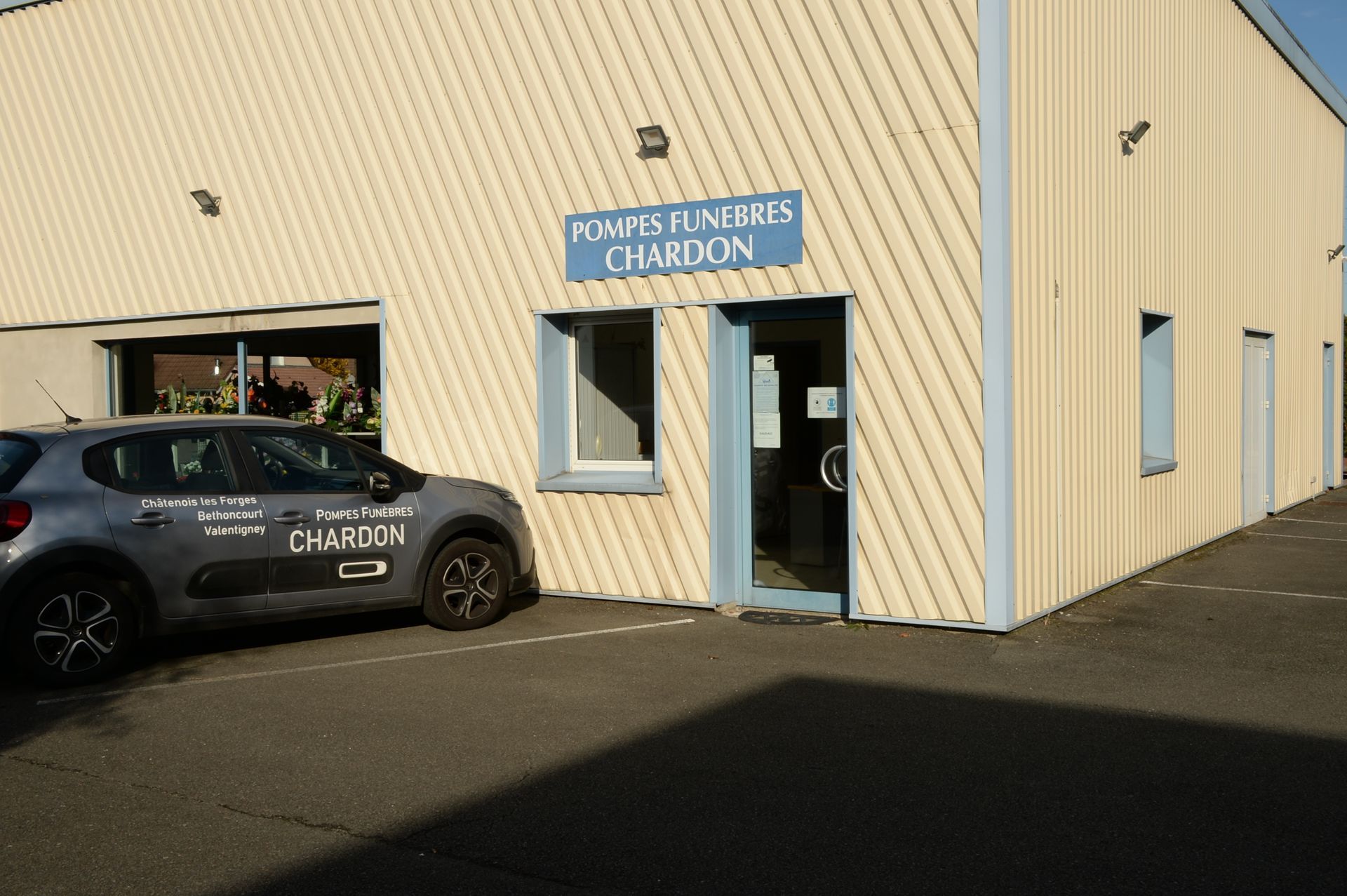 Extérieur d'un bâtiment : panneau Pompes Funèbres CHARDON au-dessus de la porte ; voiture garée, couleurs bleu clair et blanc.