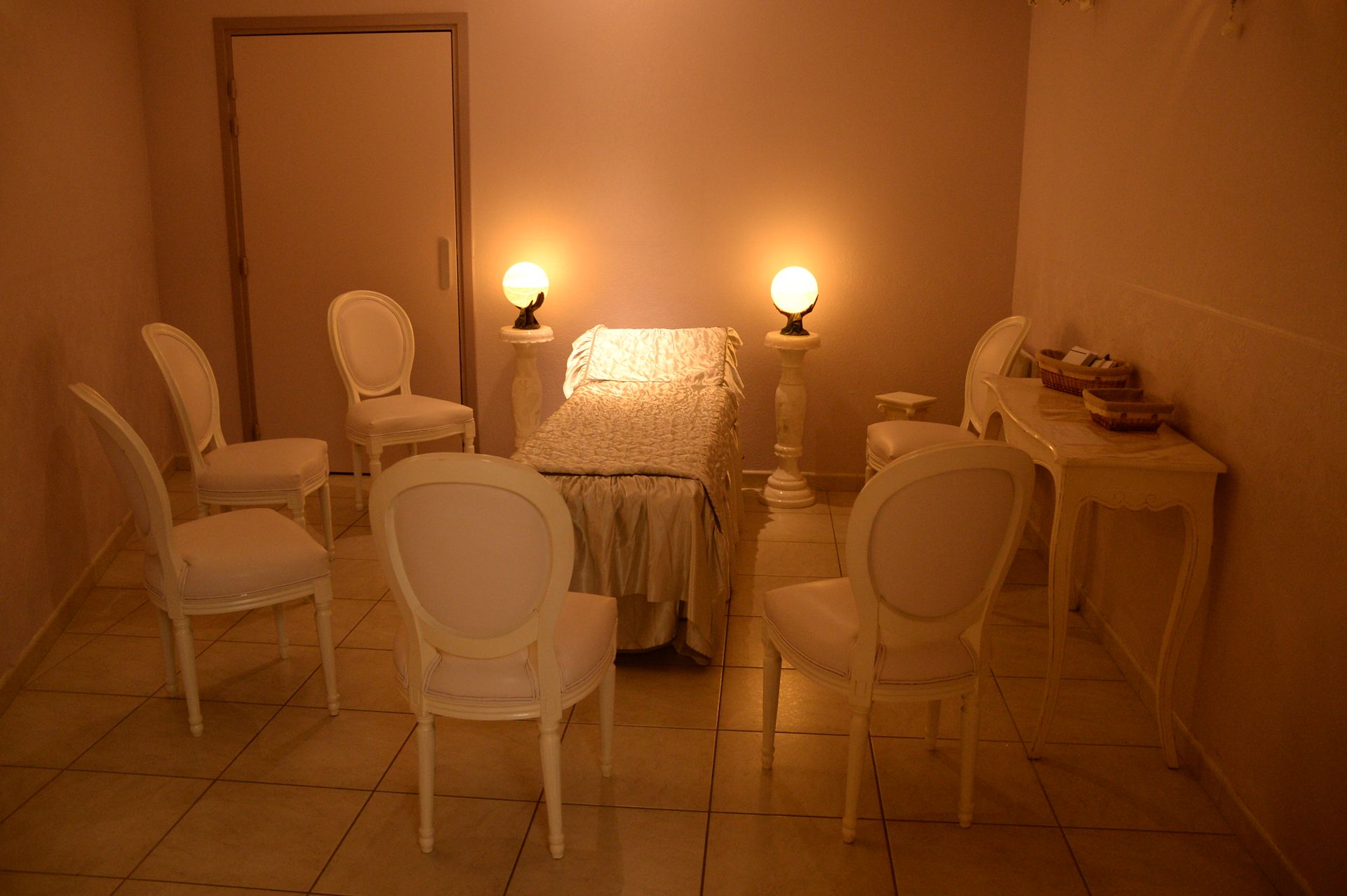 Une pièce blanche avec six chaises disposées autour d'un lit, éclairée par des lampes tamisées.