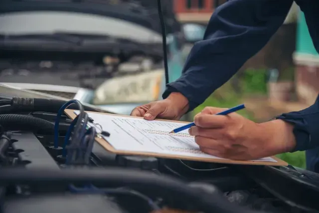 Mecánico escribiendo en un portapapeles junto a un motor de coche abierto.