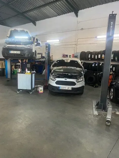Taller mecánico con dos coches en elevadores y otro en el suelo junto a pilas de neumáticos.