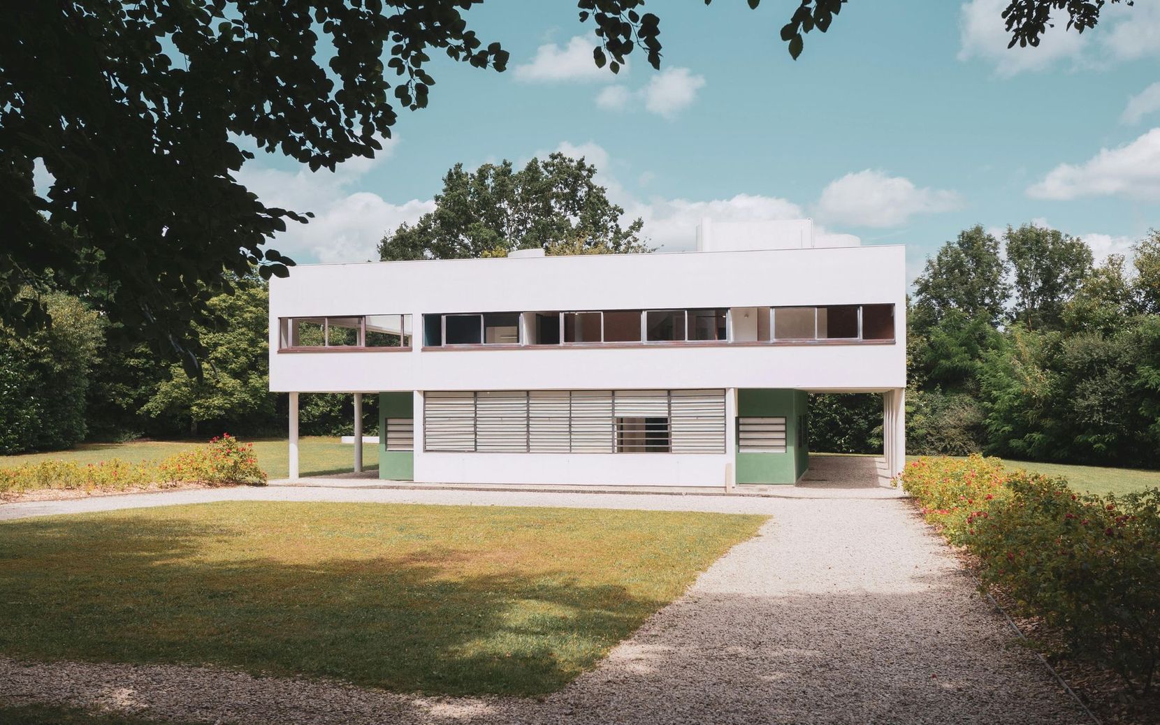 Maison moderne blanche de deux étages avec de grandes fenêtres, entourée de pelouse et d'arbres sous un ciel bleu.