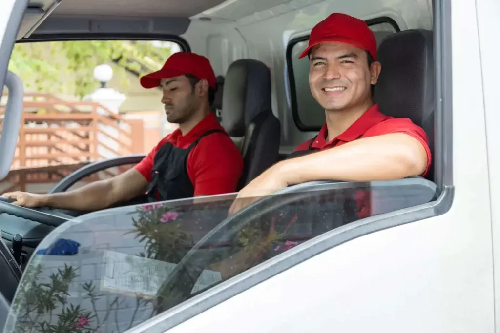 Dos repartidores con uniformes rojos y gorras en un camión, uno de ellos sonriendo a la cámara.
