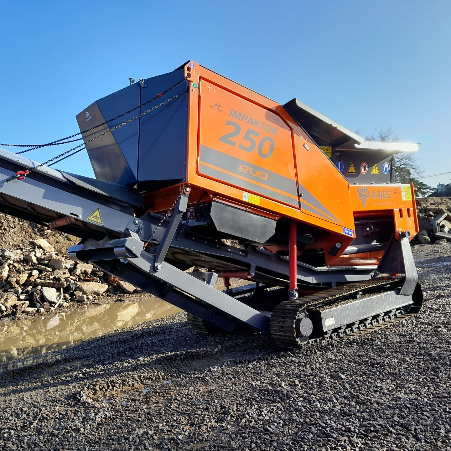 Concasseur de roches mobile orange et gris sur une surface de gravier, à l'extérieur, en plein jour.