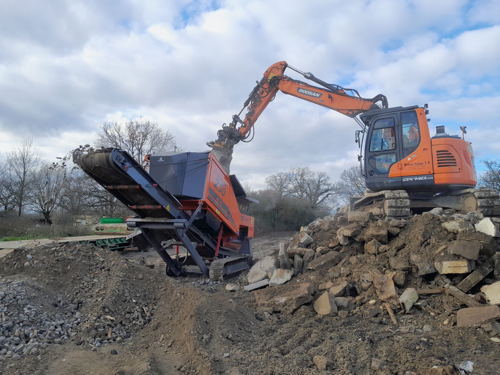 Une excavatrice orange charge des débris dans un concasseur mobile sur un chantier de construction.