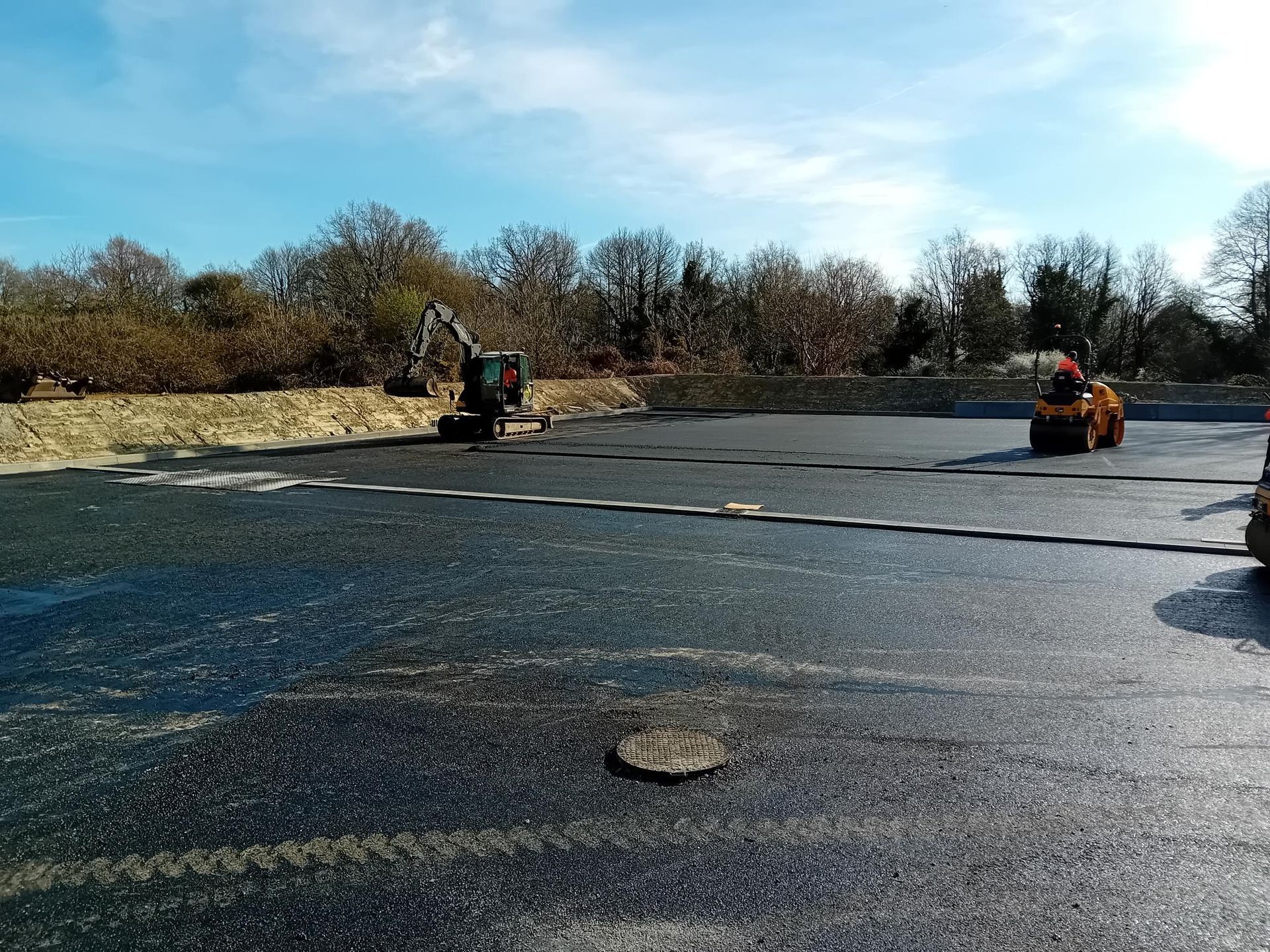 Projet de pavage d'asphalte en cours ; excavatrice, rouleau compresseur et ouvriers sur la surface nouvellement posée sous un ciel bleu.
