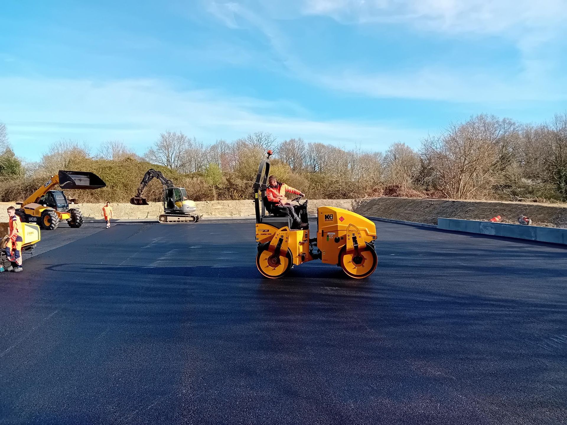 Un rouleau compresseur jaune compacte de l'asphalte noir frais. Chantier avec d'autres engins et des arbres.