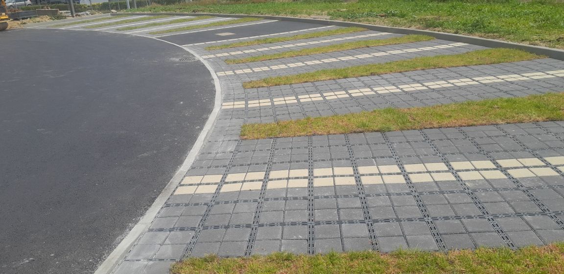 Route asphaltée sinueuse avec pavés perméables, alternant avec des bandes de gazon vert, et une zone herbeuse en arrière-plan.