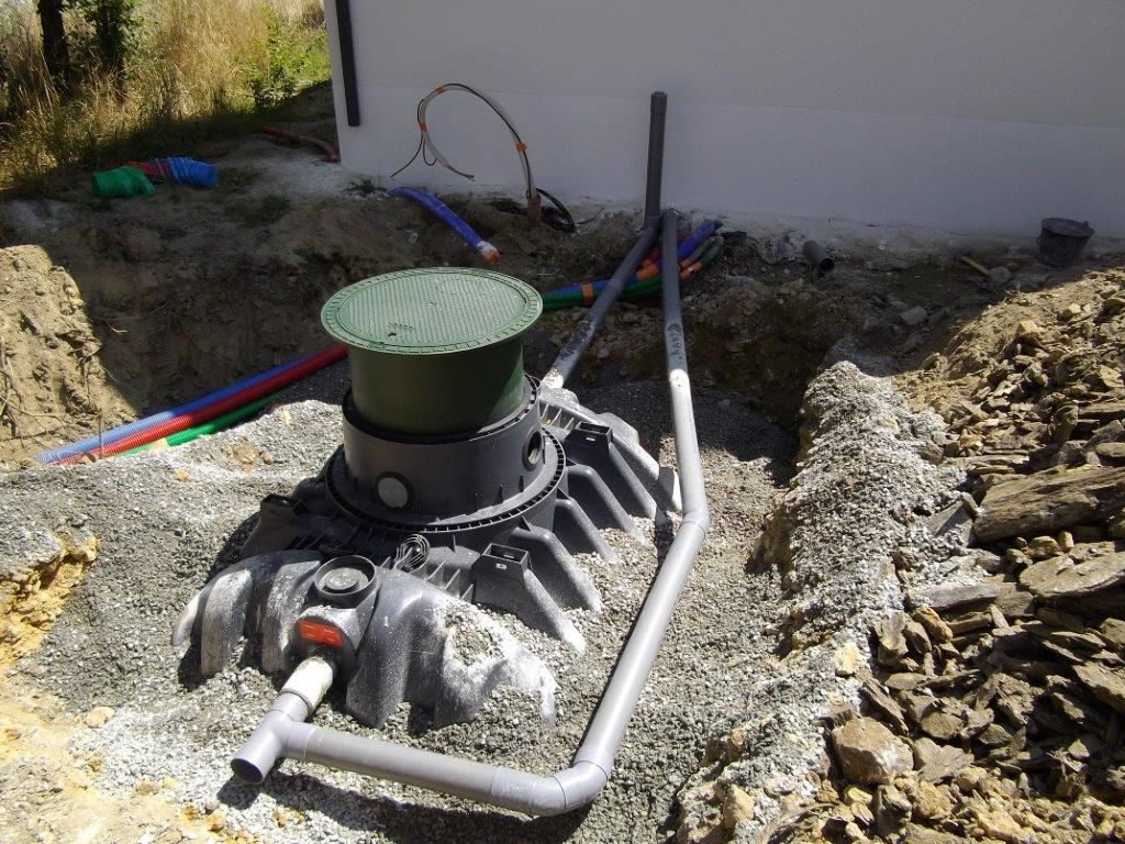 Installation d'une fosse septique en cours ; fosse grise avec couvercle vert, entourée de gravier et de tuyaux.