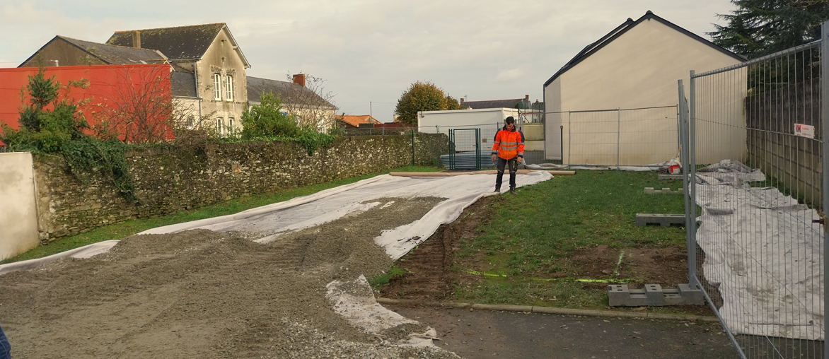 Une personne vêtue d'un gilet orange se tient devant un bâtiment blanc avec une allée et de l'herbe verte, sous un ciel gris.