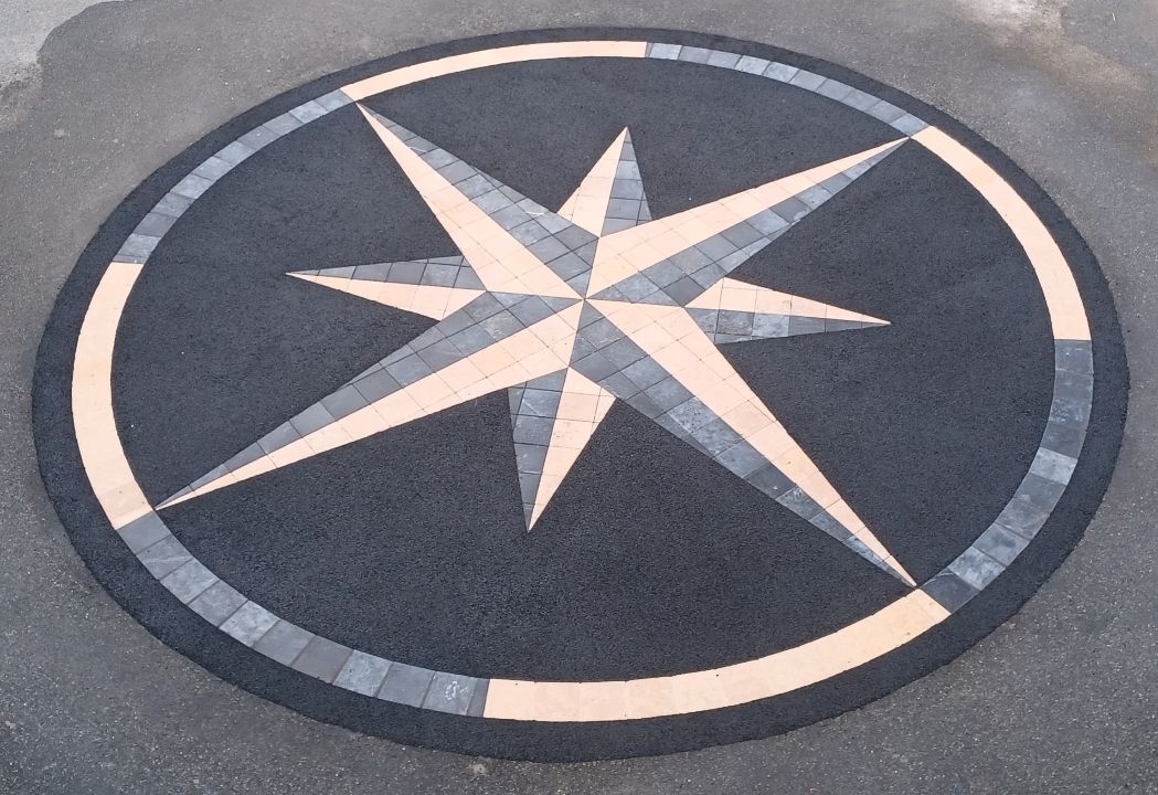 Incrustation circulaire de chaussée représentant une rose des vents dans des tons de noir, de gris et de beige.