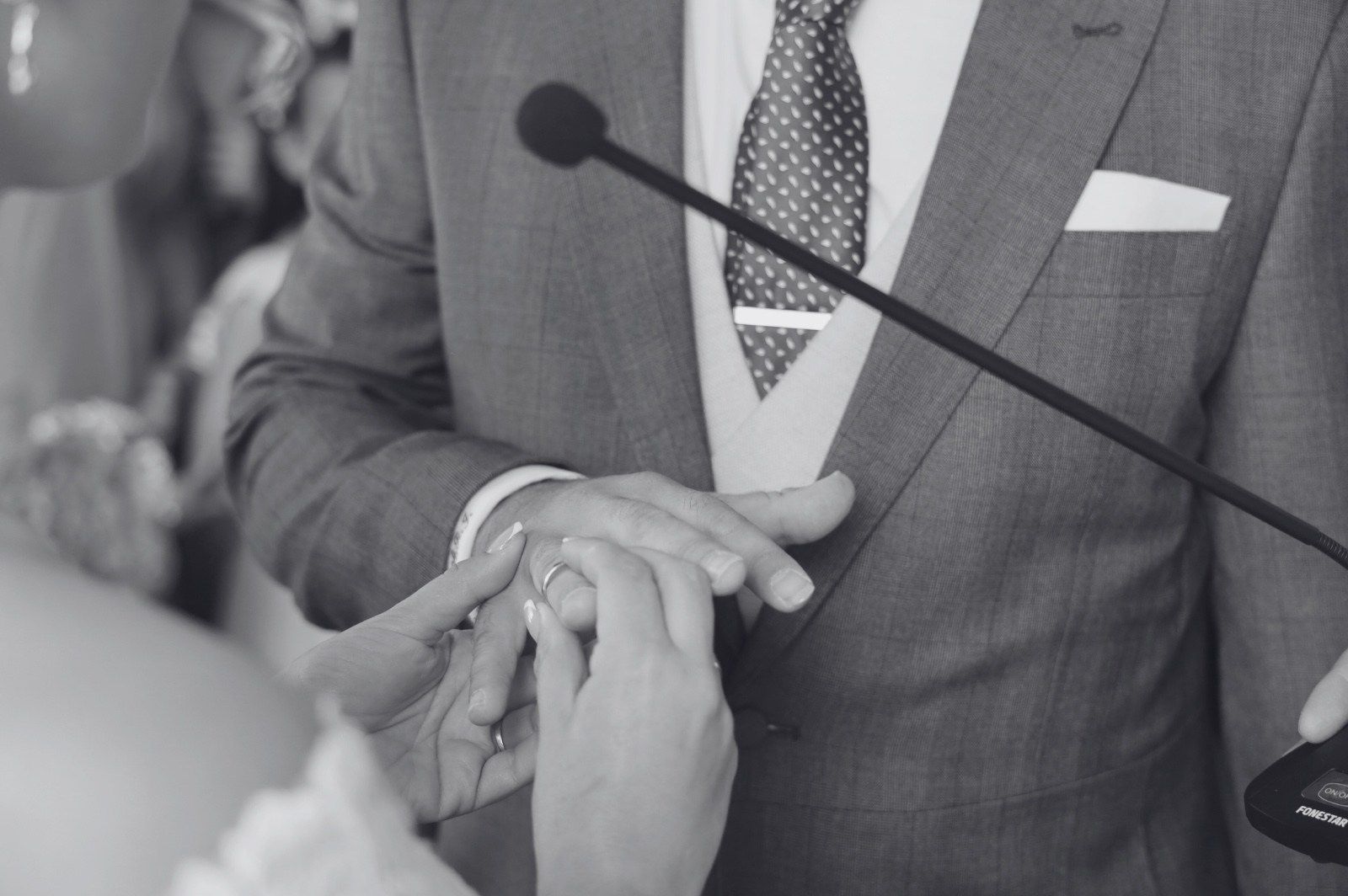 A una persona con traje se le coloca un anillo en el dedo durante una ceremonia de boda; blanco y negro.